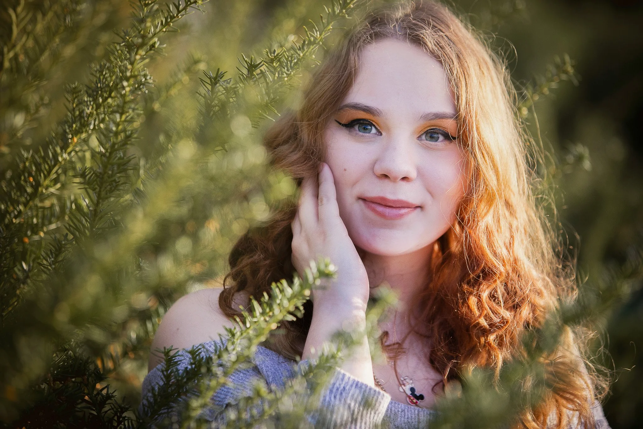 Young woman with red curly hair and blue eyes, wearing a gray off-the-shoulder top and Mickey Mouse necklace, smiling among green foliage during sunlight.