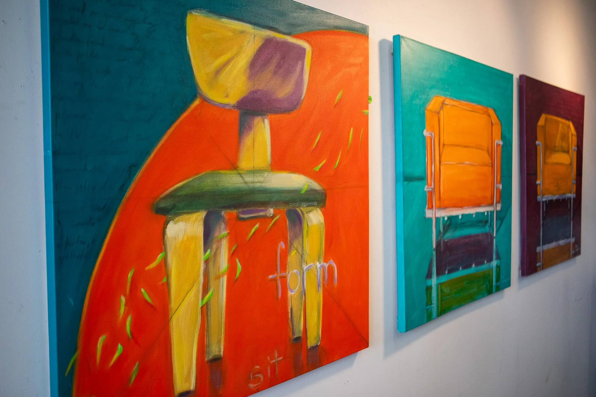 Three abstract paintings on a wall, depicting colorful chairs with vibrant backgrounds.