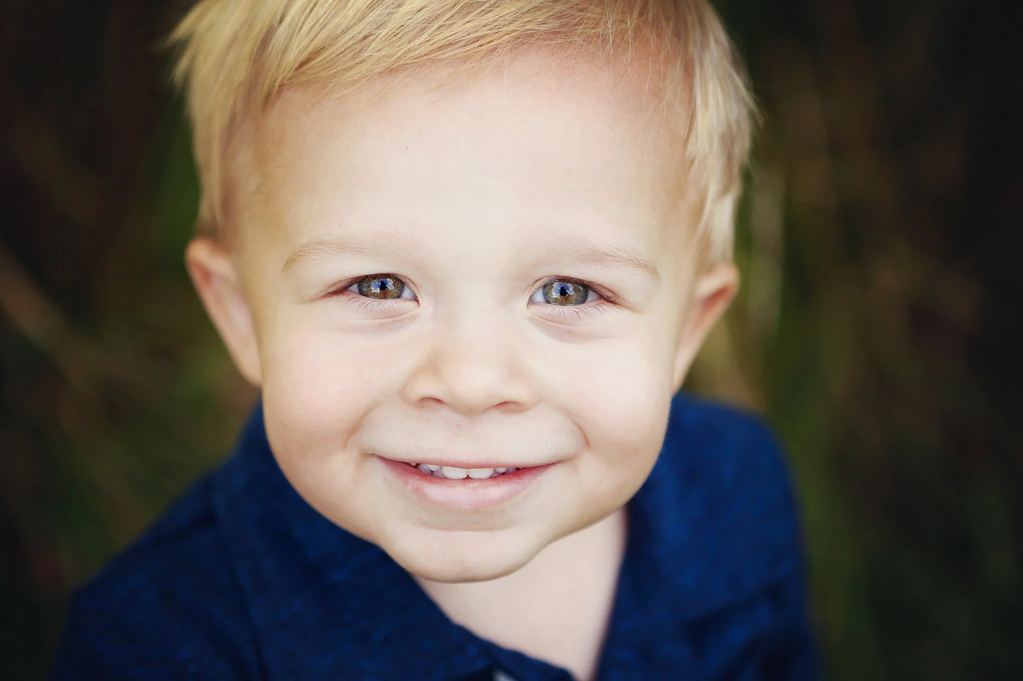 Close-up of a young boy with blonde hair and blue eyes, smiling at the camera, wearing a dark blue shirt, with a blurred outdoor background.