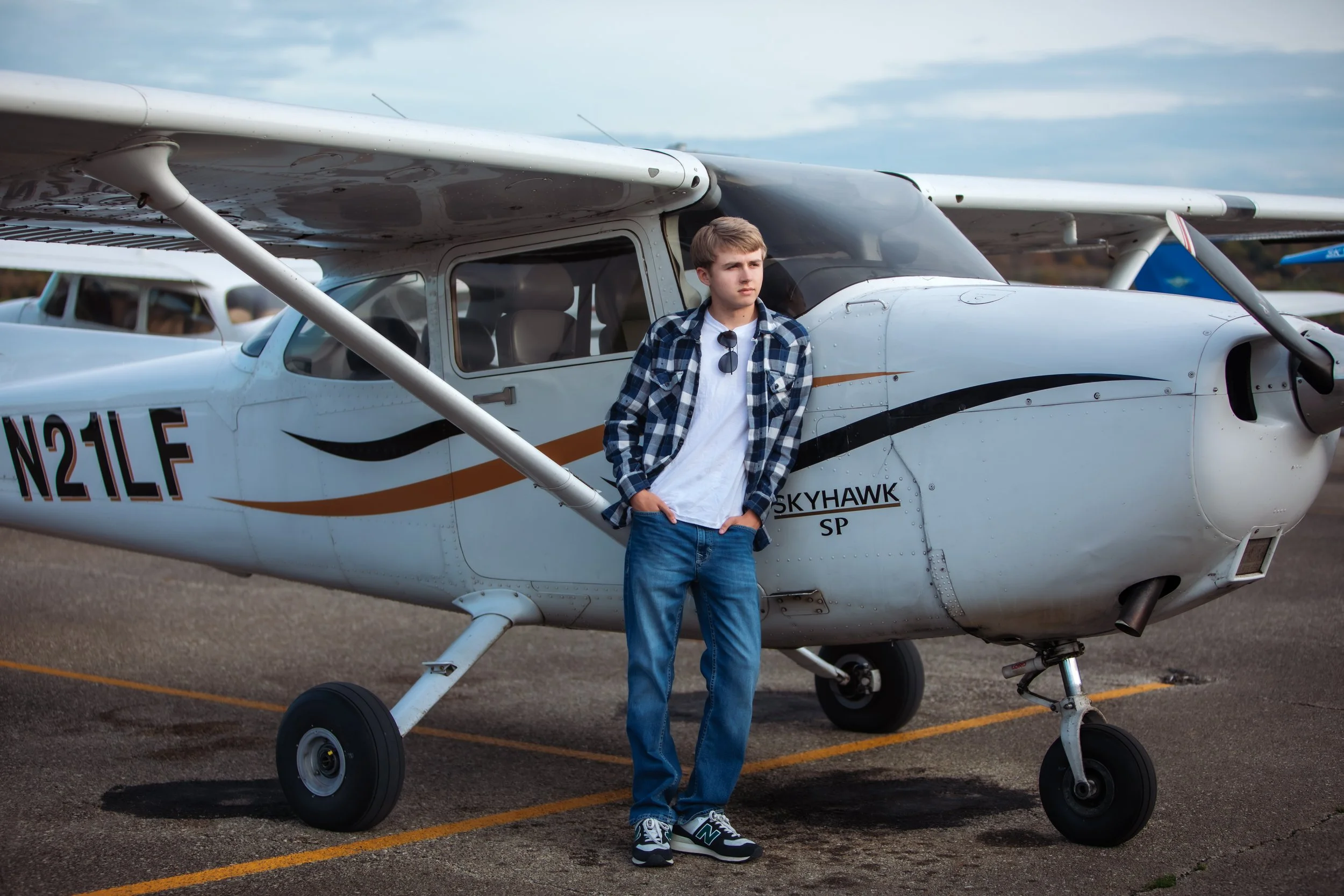 A young man standing next to a small white airplane with the word Skyhawk on the side, parked on a tarmac with a cloudy sky in the background.