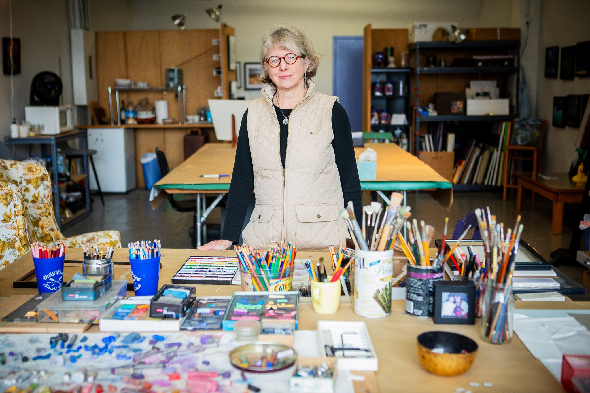 A woman with blonde hair, glasses, and a beige vest standing behind a table filled with art supplies in an art studio.
