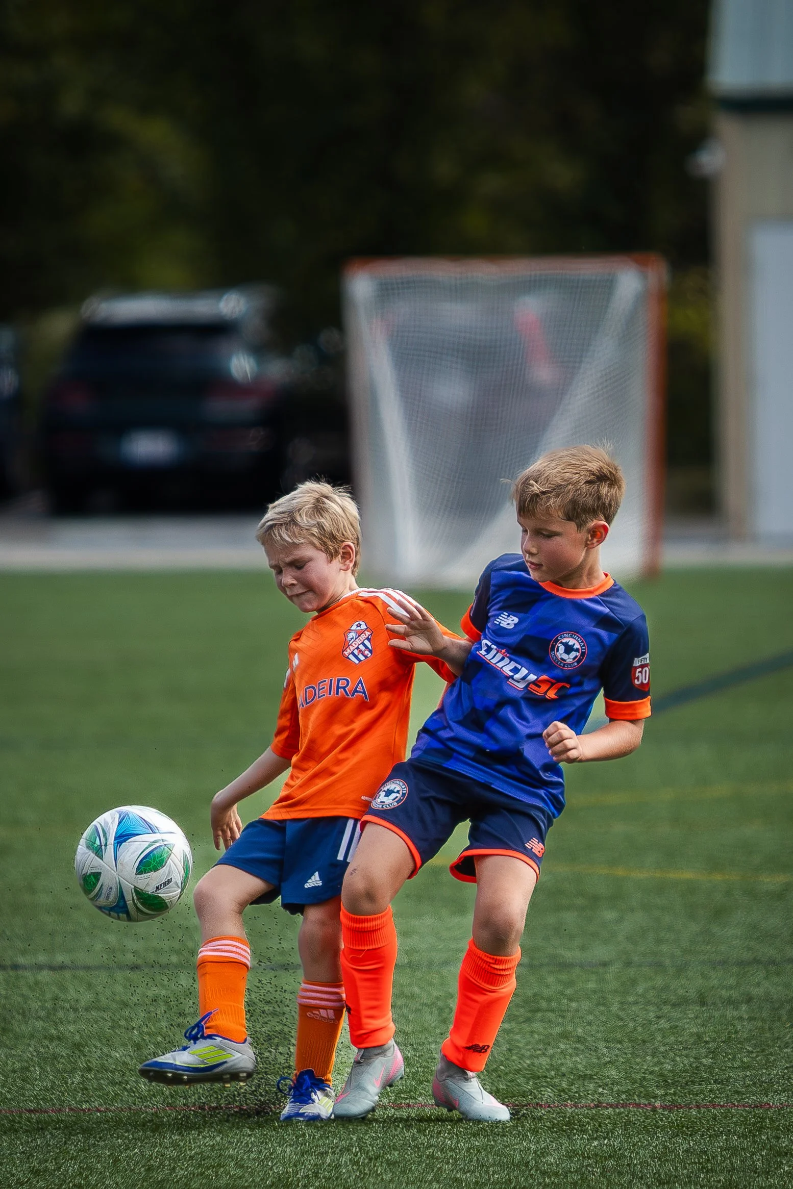 Two boys playing soccer on a field, one in an orange jersey and the other in a blue jersey, competing for the ball.