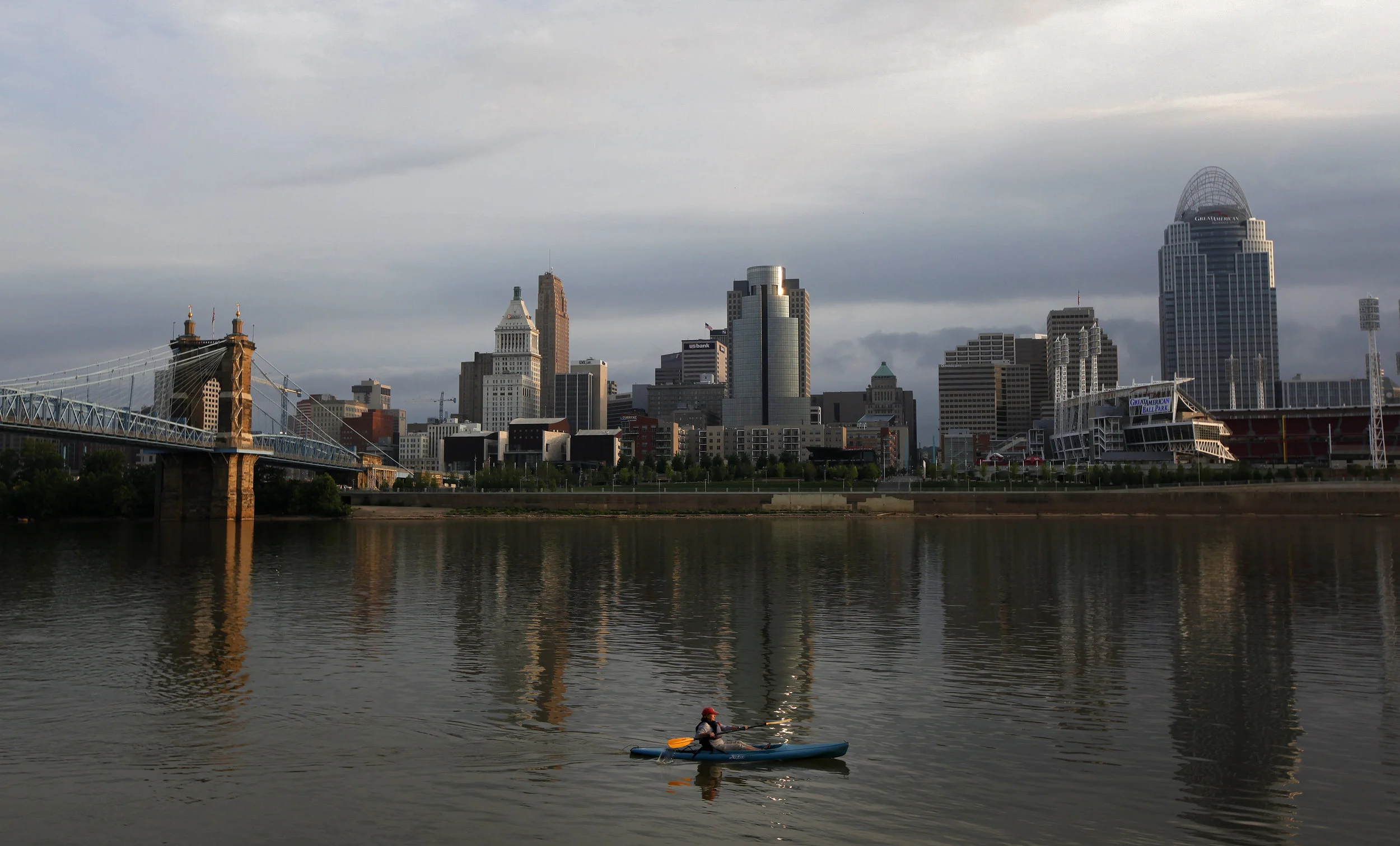 A person kayaking on a river with a city skyline and bridge in the background, under a cloudy sky.
