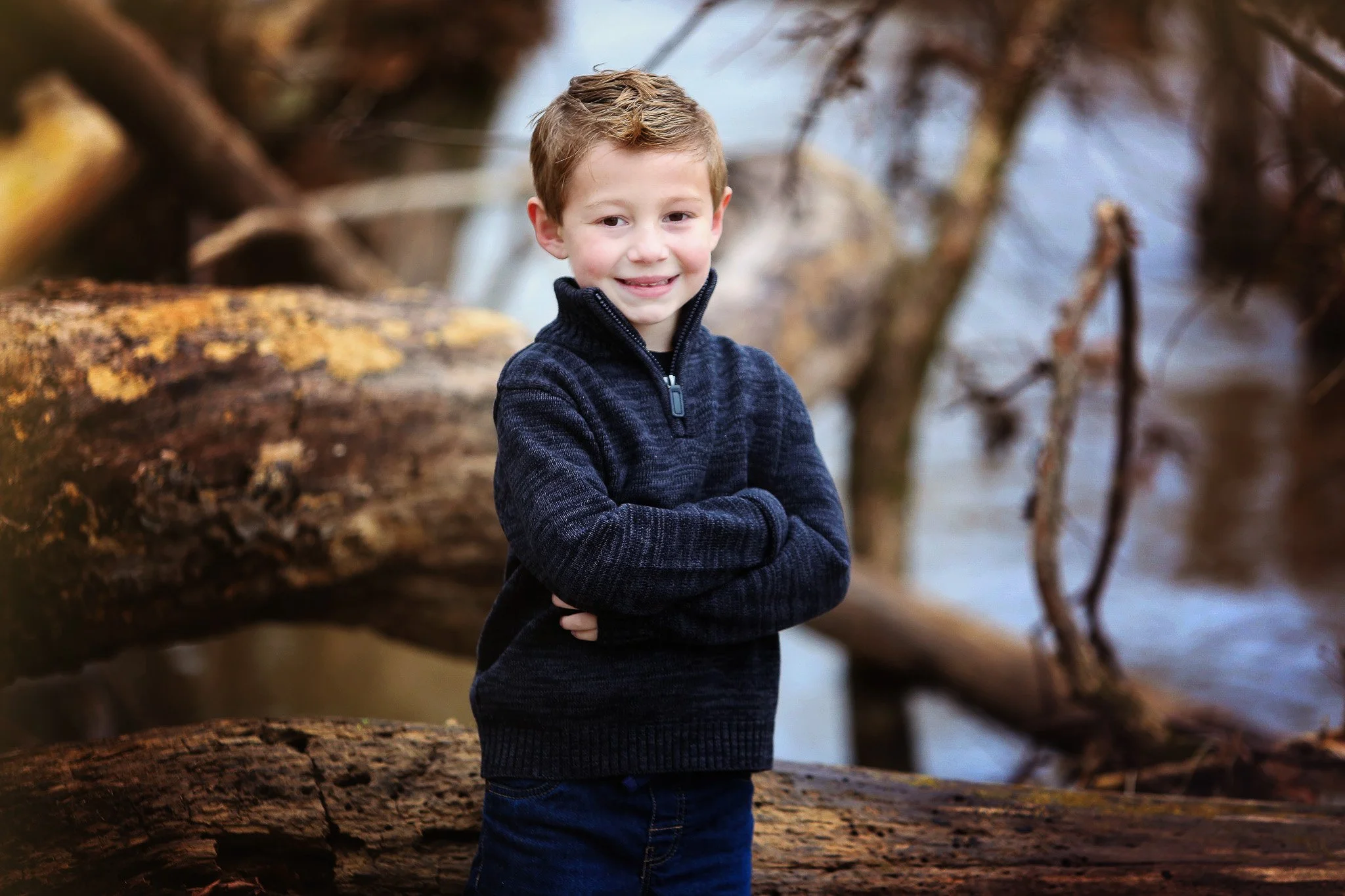 A young boy with blonde hair, wearing a dark blue sweater with a zip collar, standing outdoors by a river with fallen logs and trees in the background, smiling with arms crossed.