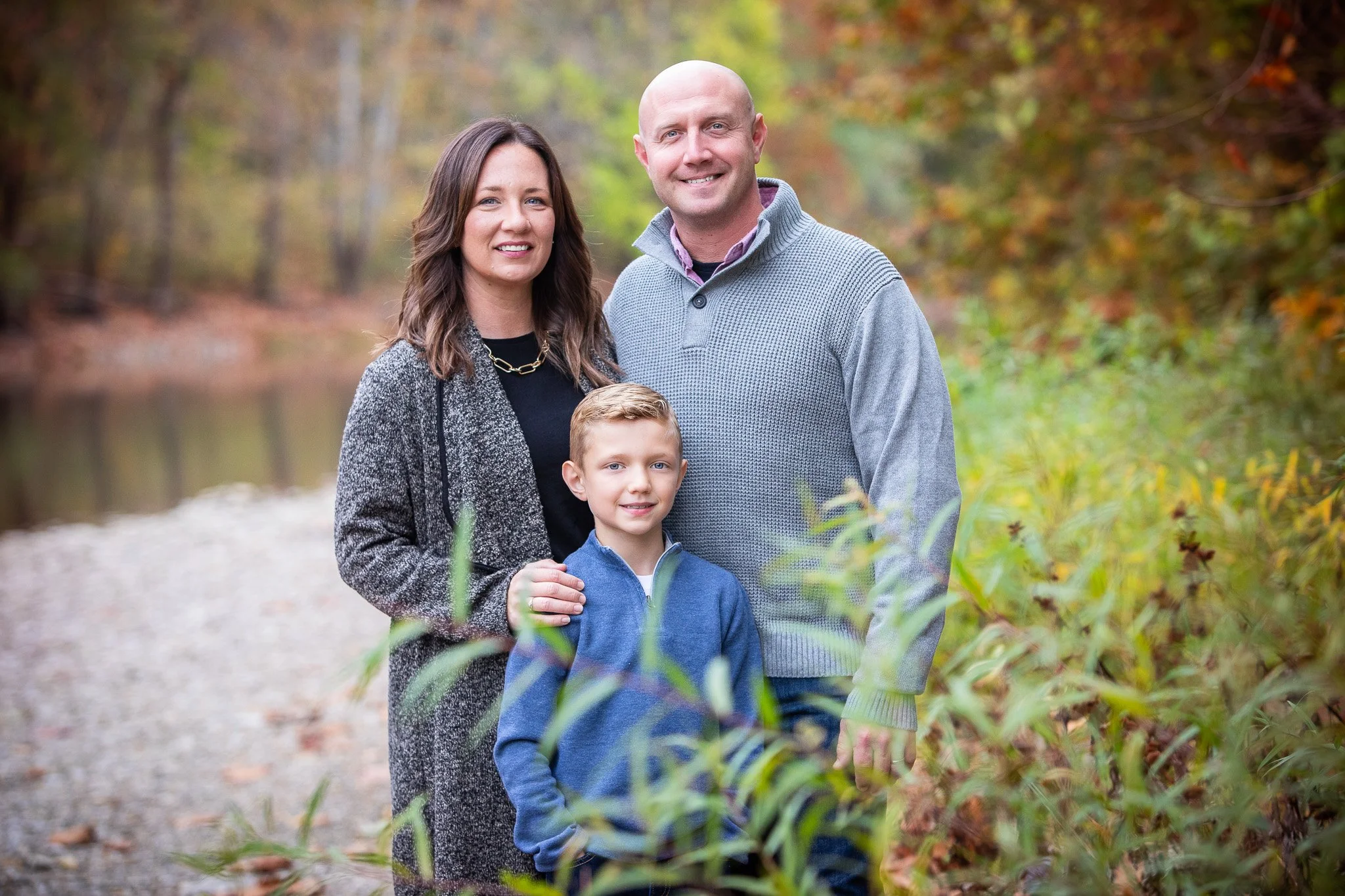 Family of three standing outdoors near a river with autumn foliage in the background. The woman has shoulder-length brown hair, the man is bald, and the boy has short blonde hair. They are dressed in casual fall clothing.