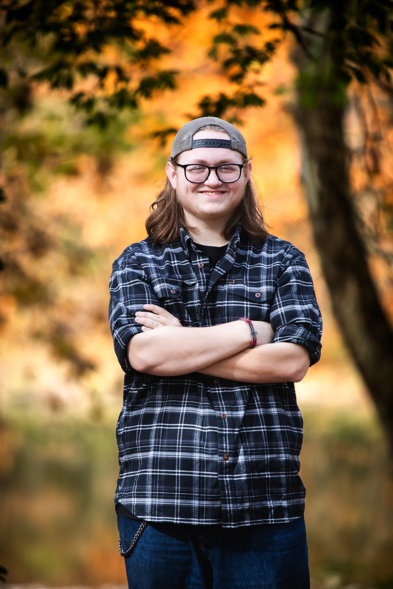 A young man with long hair, glasses, and a gray cap, smiling with his arms crossed, standing outdoors in a forest during fall with orange and yellow leaves.