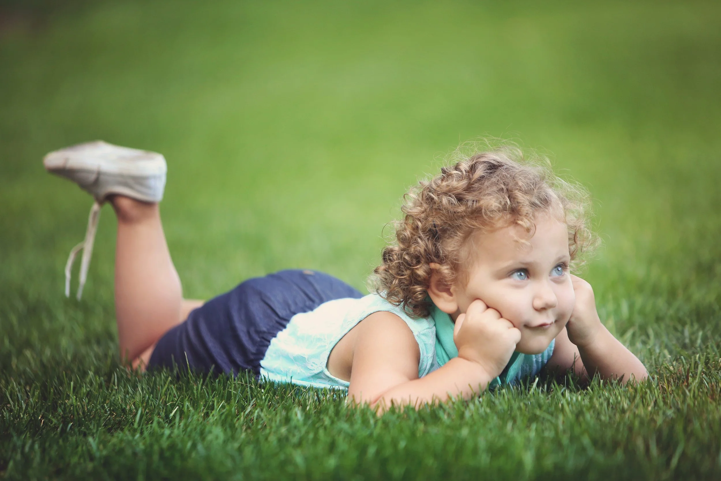 Young child with curly hair lying on grass, resting chin on hands, looking off to the side.