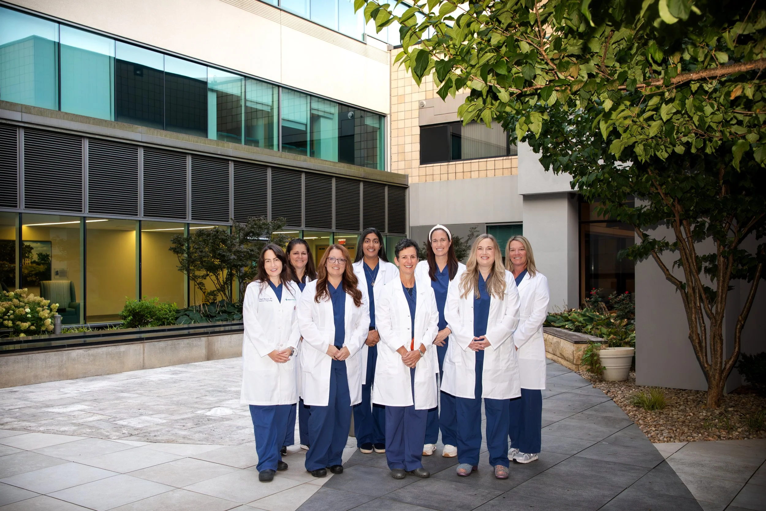 Group of nine female healthcare professionals standing outdoors in front of a modern building, wearing white lab coats and navy blue scrubs, smiling for the camera.