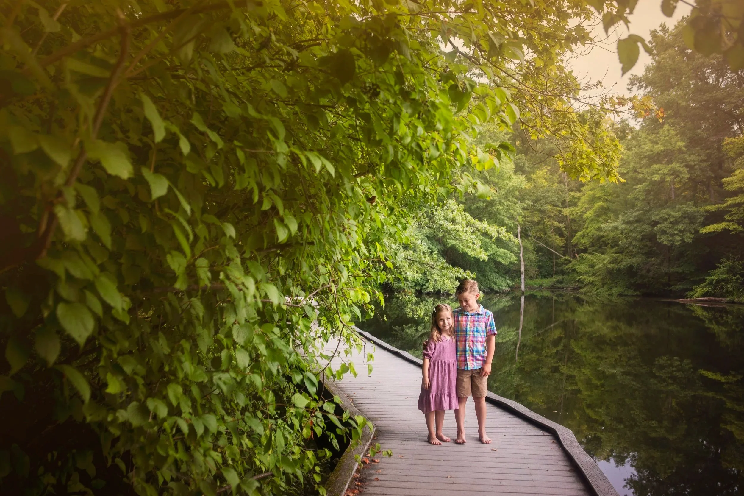 Two children, a girl and a boy, standing on a wooden dock next to a peaceful lake, surrounded by lush green trees, with the girl wearing a pink dress and the boy in a colorful plaid shirt and shorts. The scene is calm and natural, with reflections of