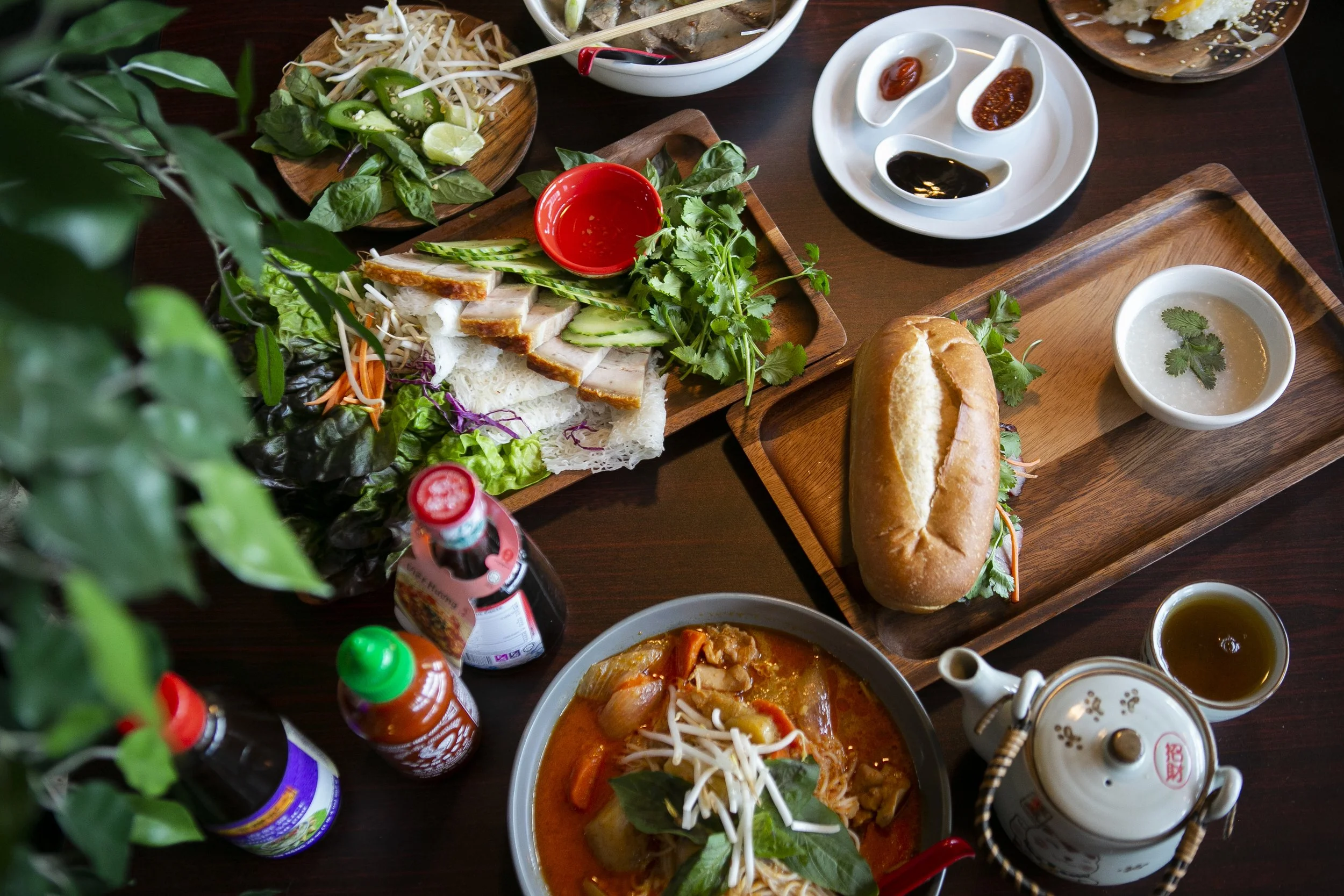A variety of Asian dishes on a dark wooden table, including a bowl of curry, a sandwich baguette, a plate of sliced pork with vegetables and herbs, and an assortment of sauces, soups, and condiments.