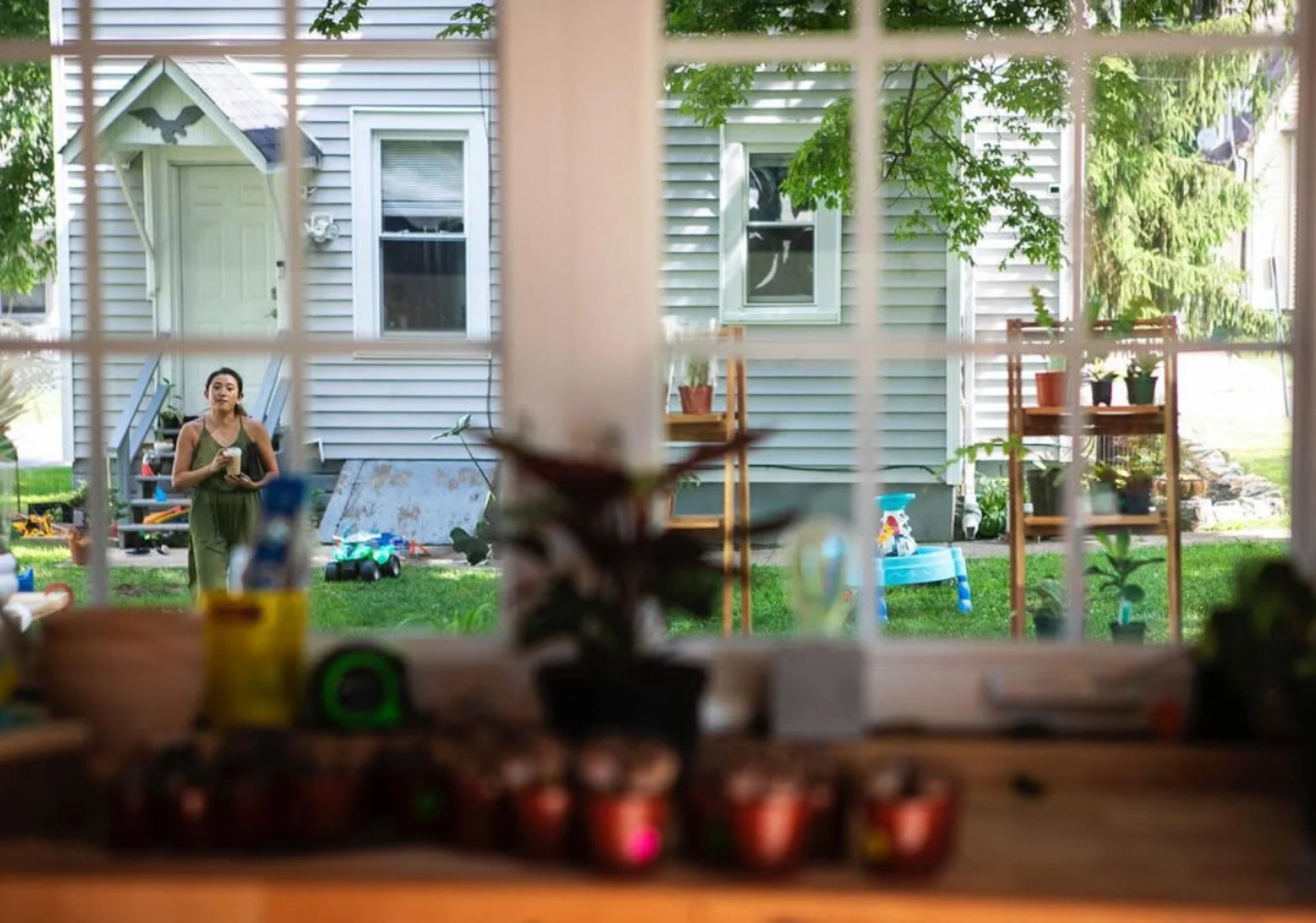 View through a window showing a woman with dark hair in a ponytail, standing outside in a grassy backyard, holding a small object, with trees and a white house in the background.
