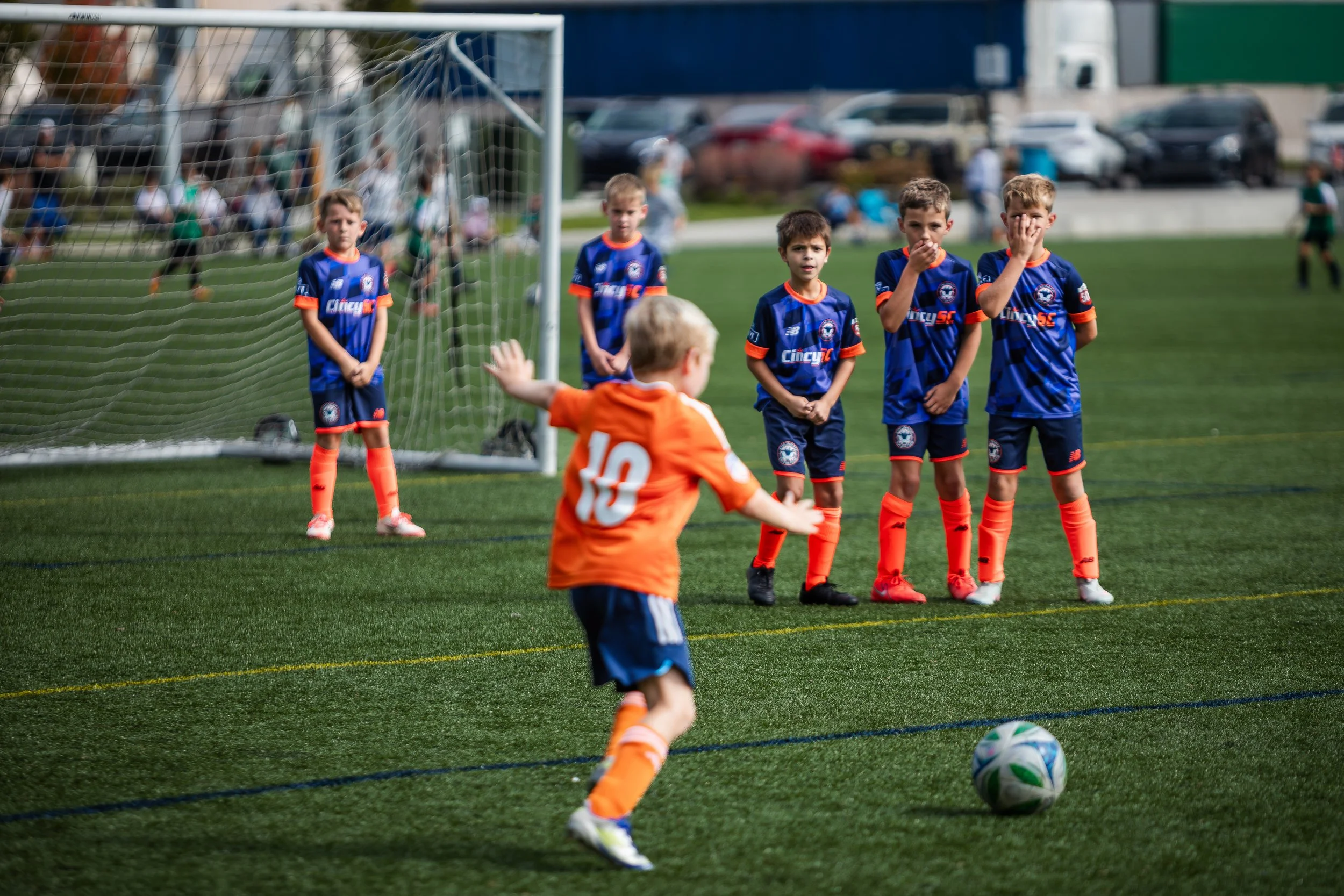 Young boy in orange soccer uniform taking a penalty shot from a soccer ball, while four boys in blue and orange uniforms stand in front of a goal, watching and preparing to block the shot, on a soccer field.