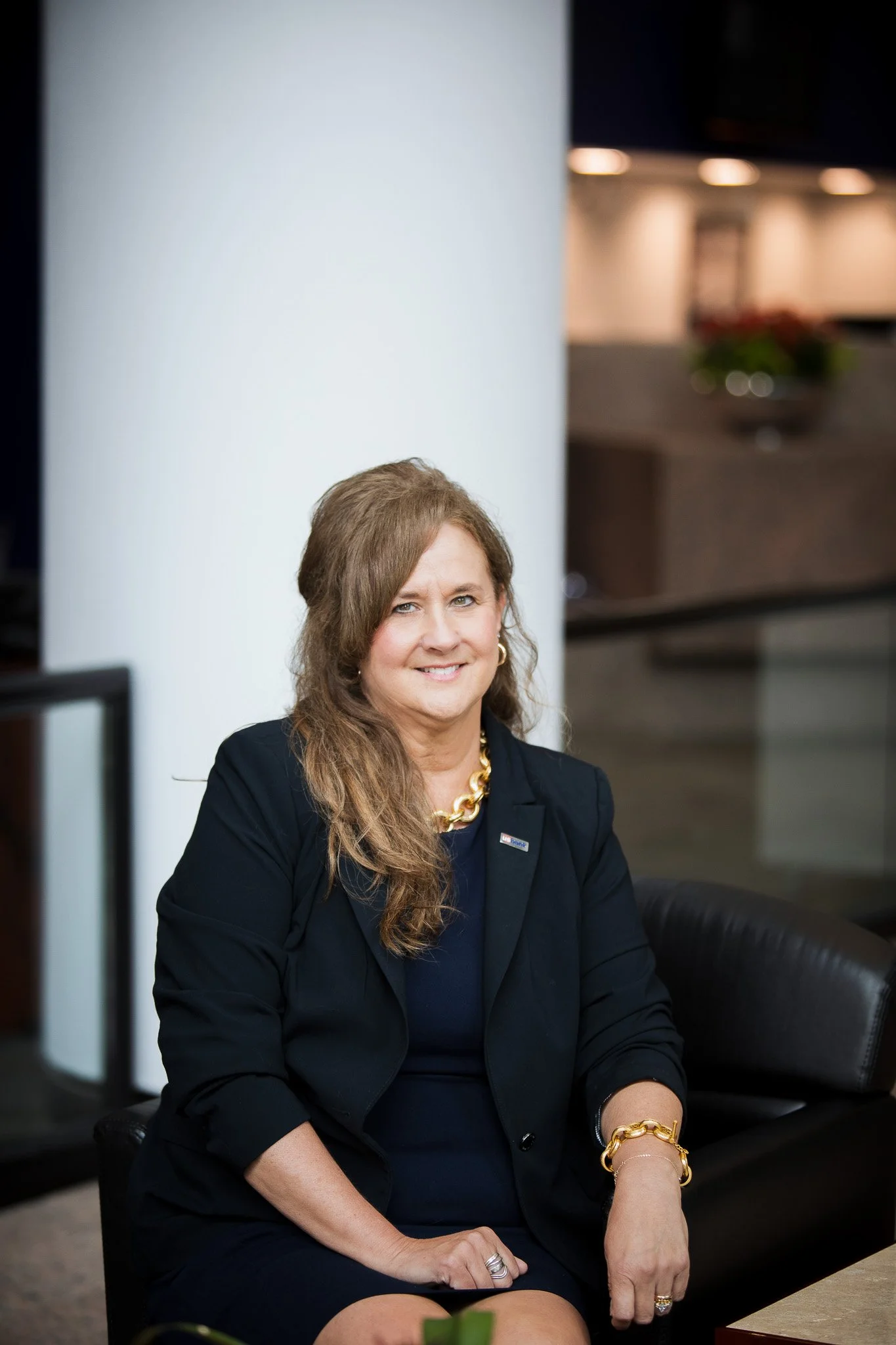 A woman with long, wavy brown hair sitting on a black leather chair, wearing a dark blazer, gold jewelry, and smiling in an indoor setting with a white pillar and blurred background.