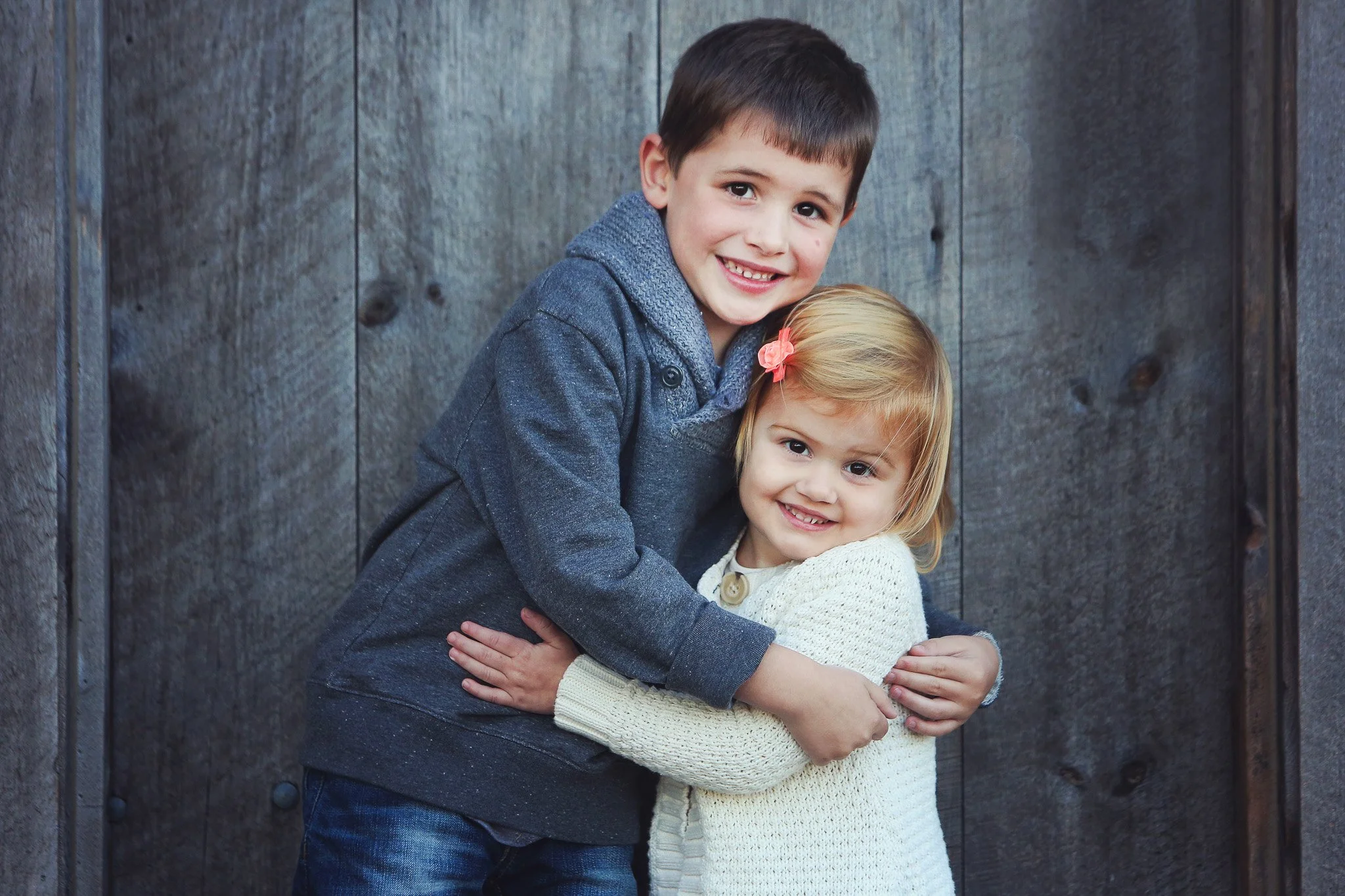 A young boy and girl hugging and smiling at a camera, standing against a wooden background.