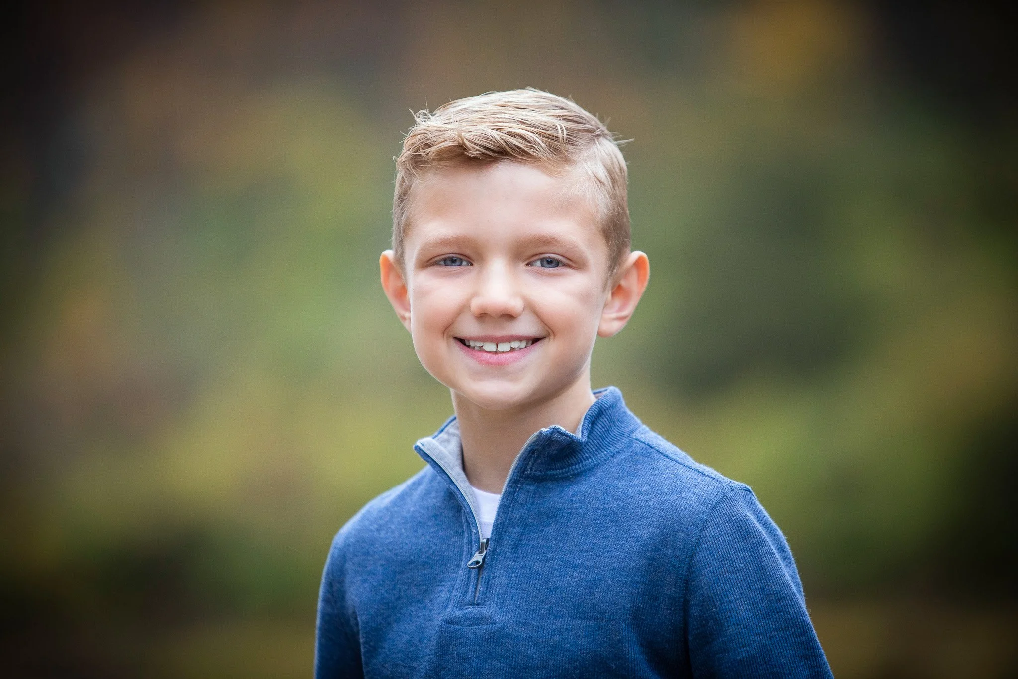 A young boy with short, light brown hair and blue eyes, smiling, outdoors with a blurred green and brown background, wearing a blue quarter-zip sweater.