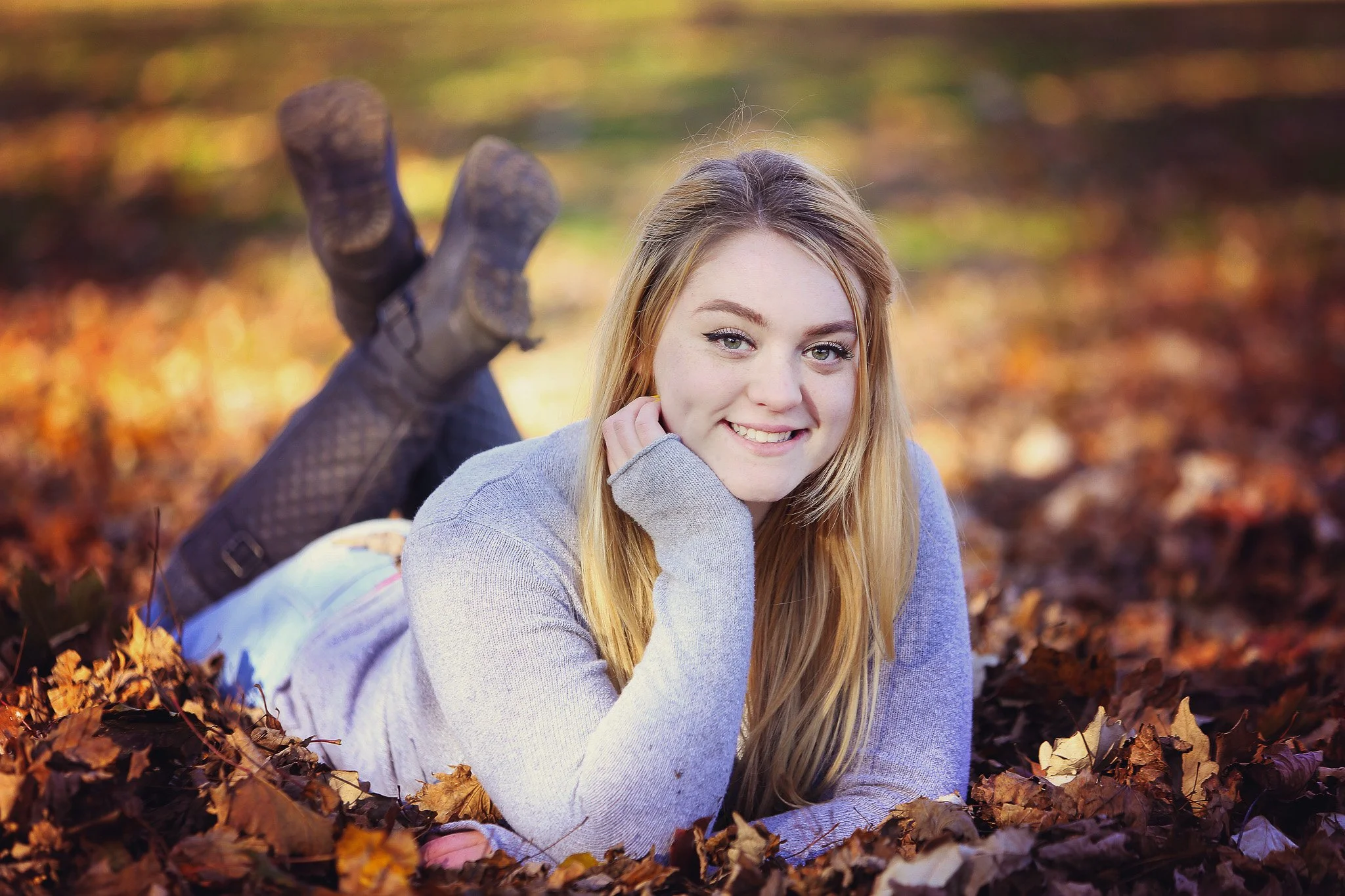 Young woman with long blonde hair lying on her stomach in a pile of fallen autumn leaves in a park, wearing a light gray sweater and black boots, smiling at the camera.