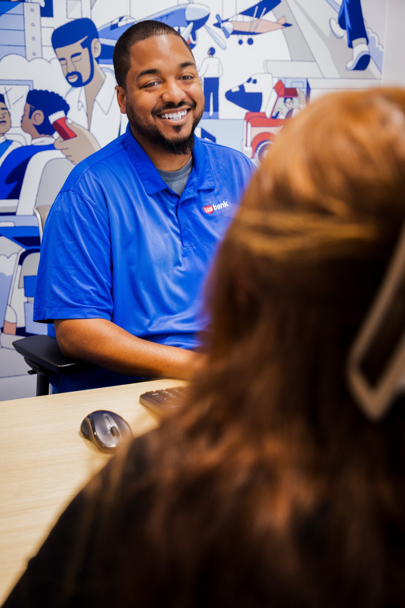 A man in a blue US Bank shirt smiling while talking to a woman with long brown hair in an office setting with a colorful mural on the wall behind them.