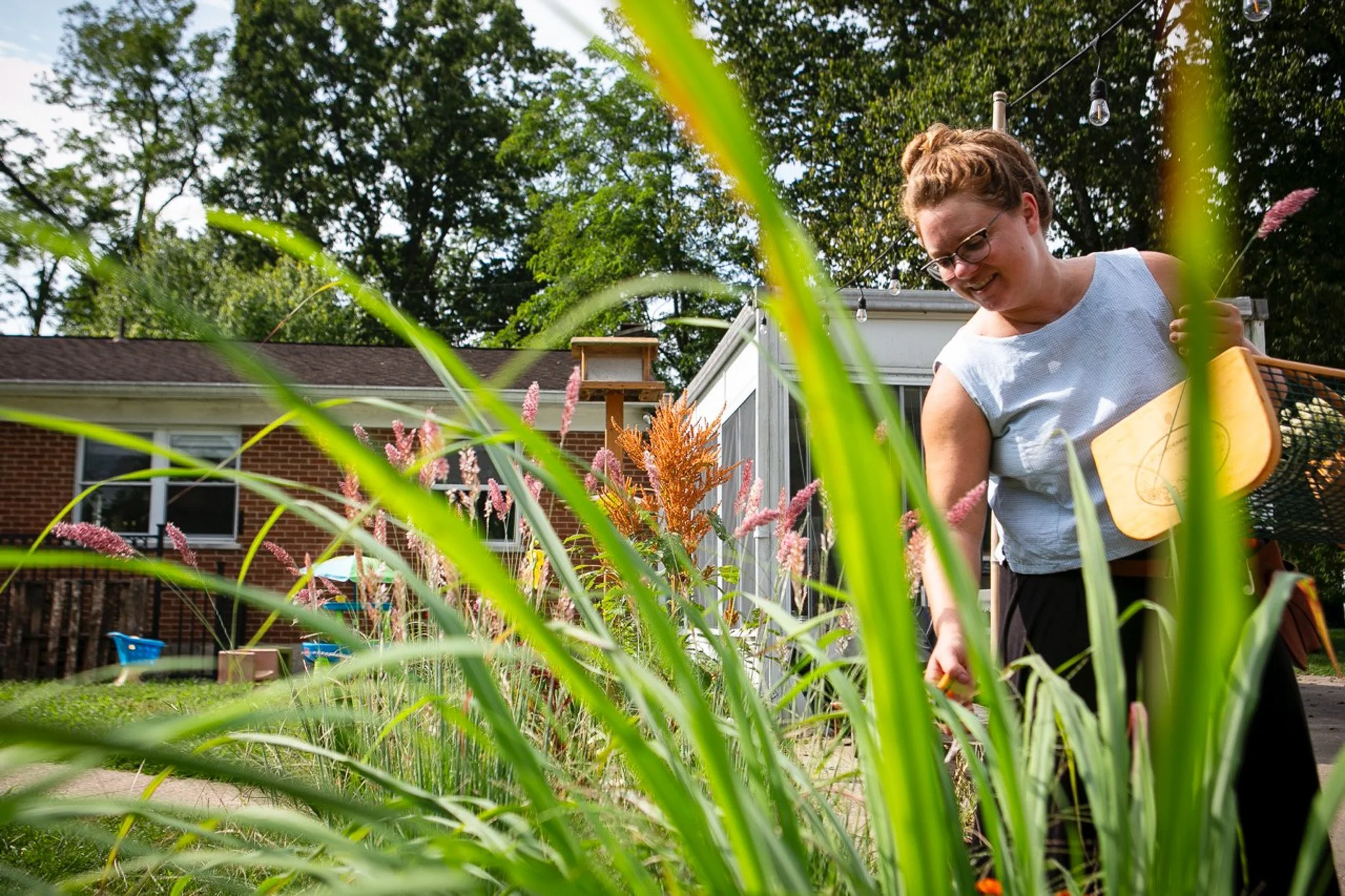 A woman gardening in a backyard with tall pink and orange flowers, a brick house, and a birdhouse on a pole in the background.