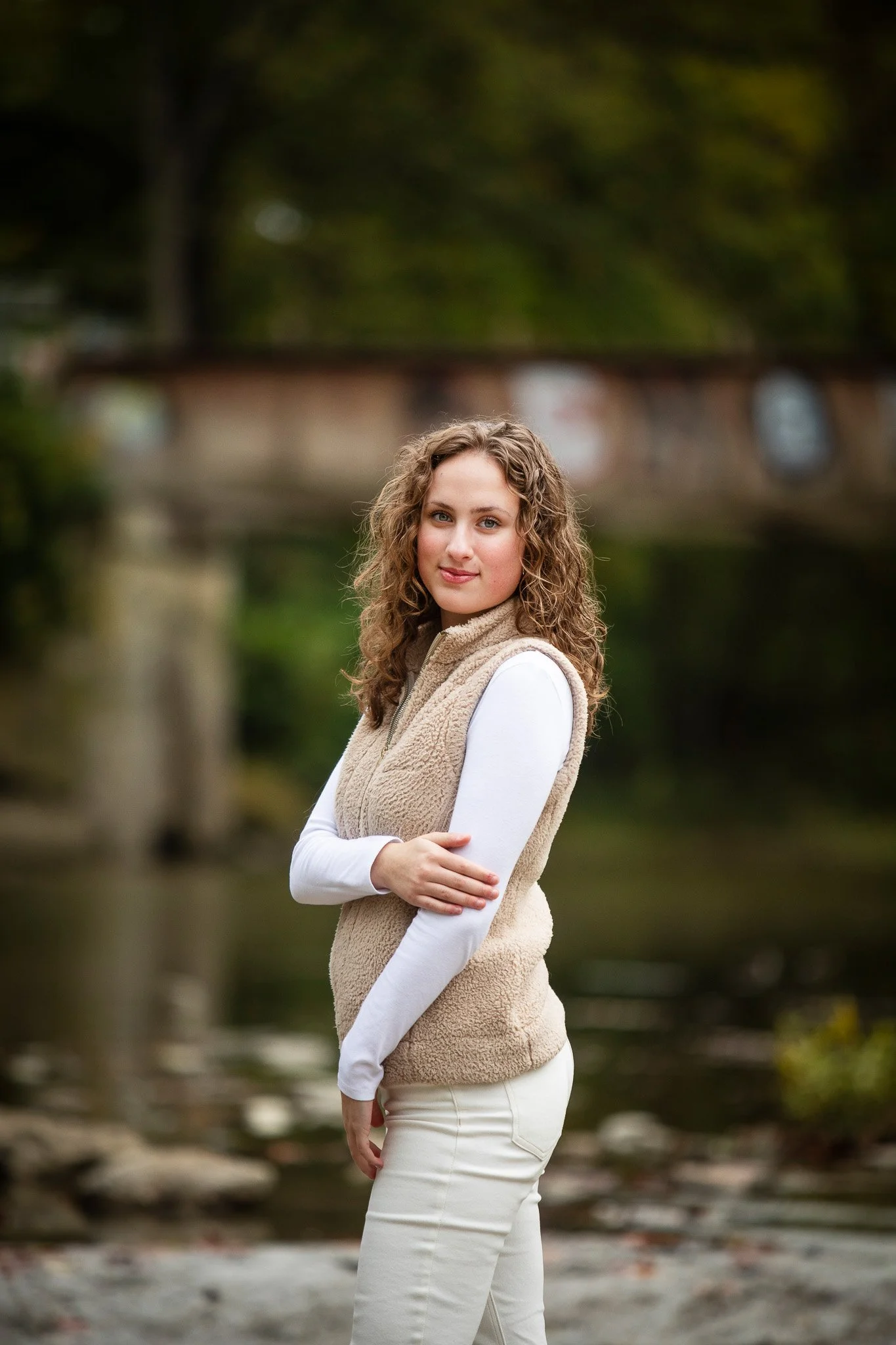 A young woman with curly hair standing outdoors by a river with a bridge in the background, wearing a beige fleece vest over a white long-sleeve shirt and cream pants.