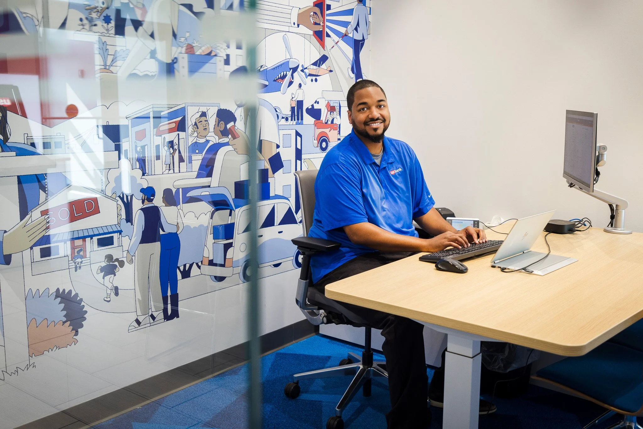 A man in a blue collared shirt sitting at a desk, smiling at the camera, in an office with a colorful mural on the wall behind him featuring illustrations of people, animals, and buildings.