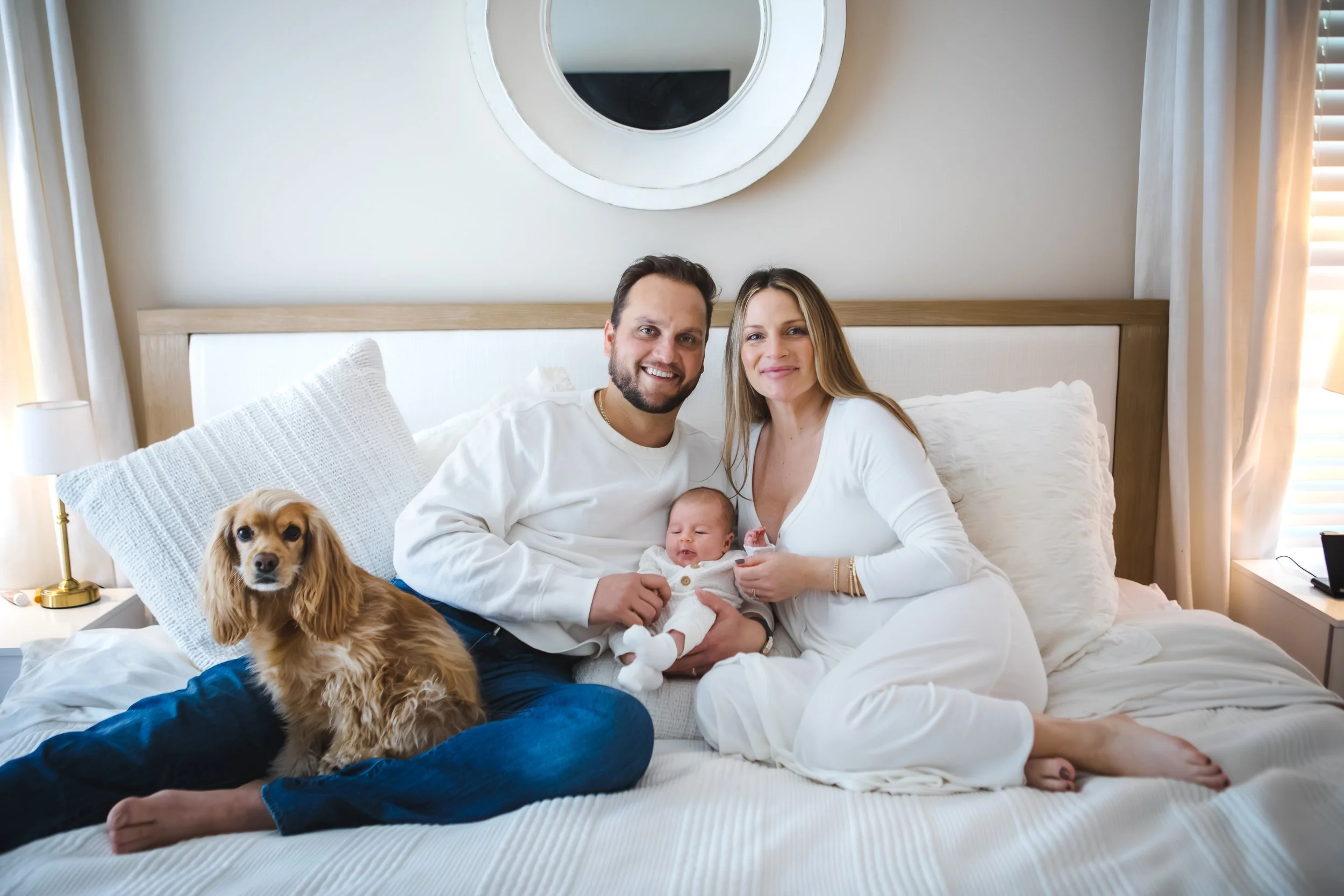 A happy family of three with a dog sitting on a bed in a bright bedroom. The father, mother, and baby are smiling. The dog is sitting on the bedding next to them.