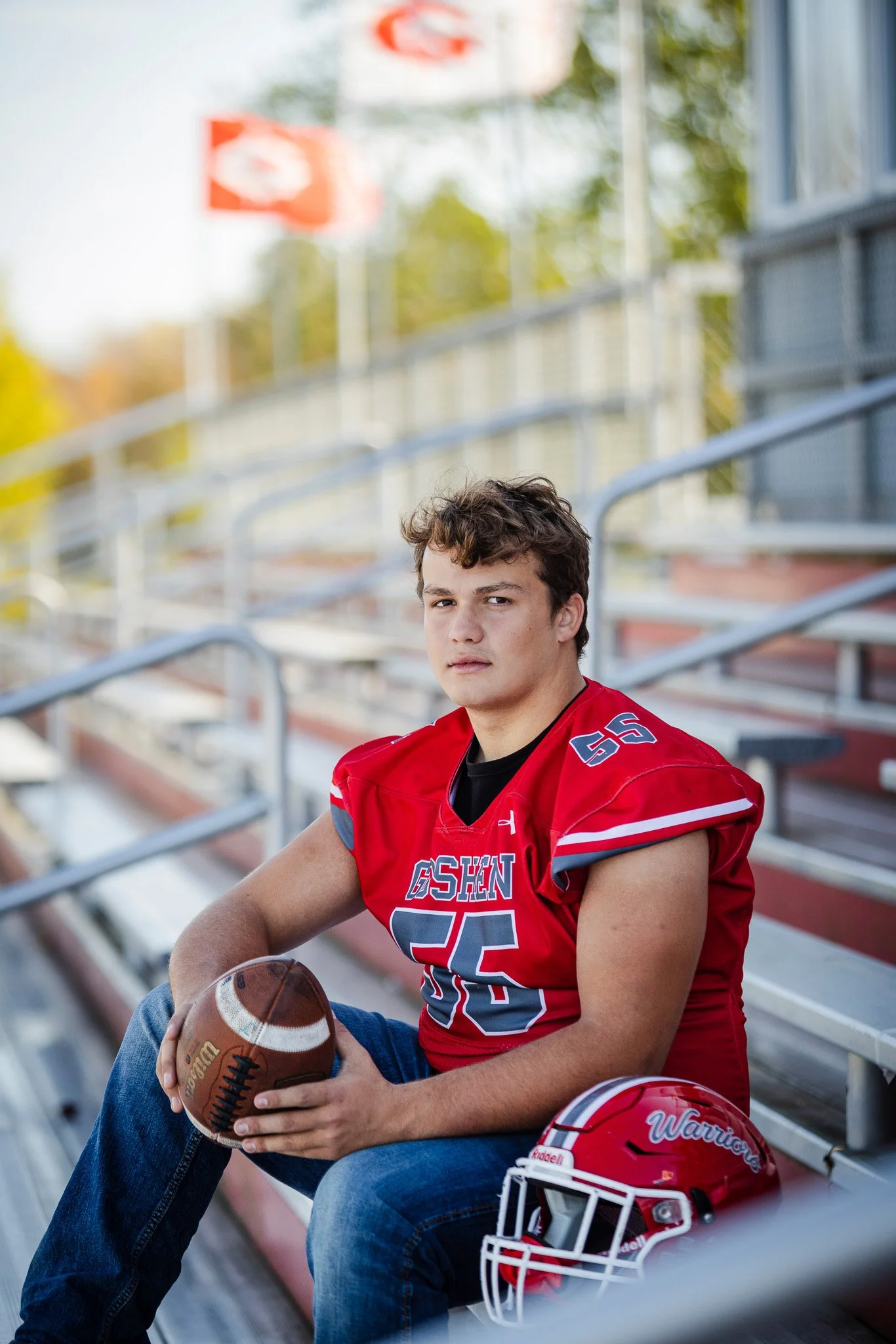 Young football player in a red uniform sitting on bleachers, holding a football, with a red football helmet on the seat next to him, in a stadium setting.
