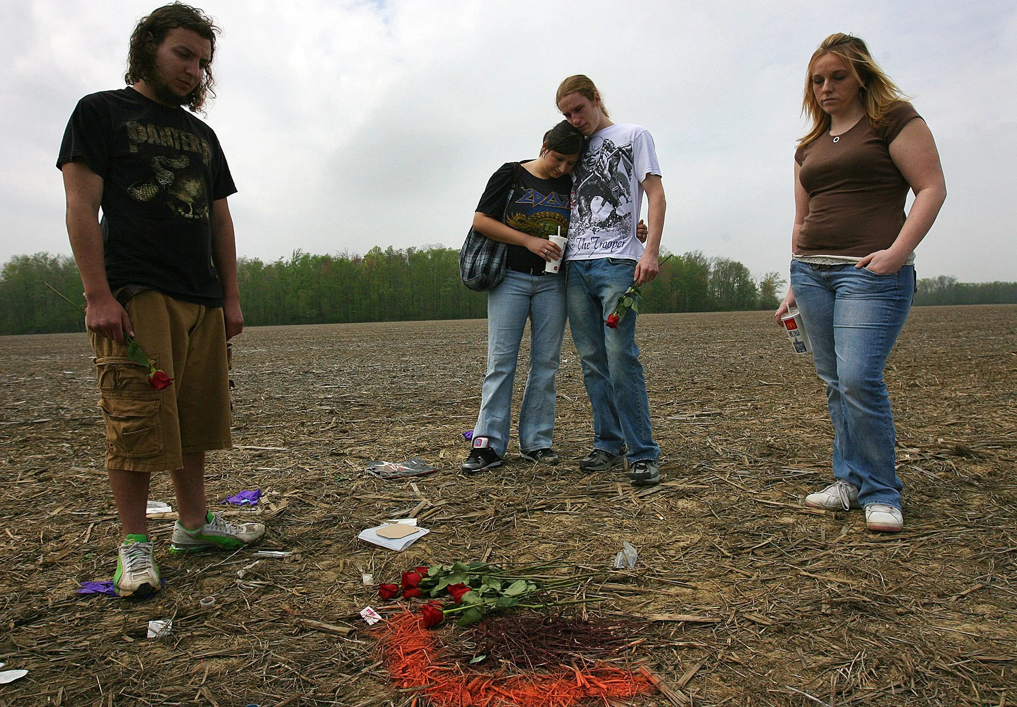 Group of four young people standing on a field during daytime, with flowers and memorial items placed on the ground in front of them, possibly commemorating someone.