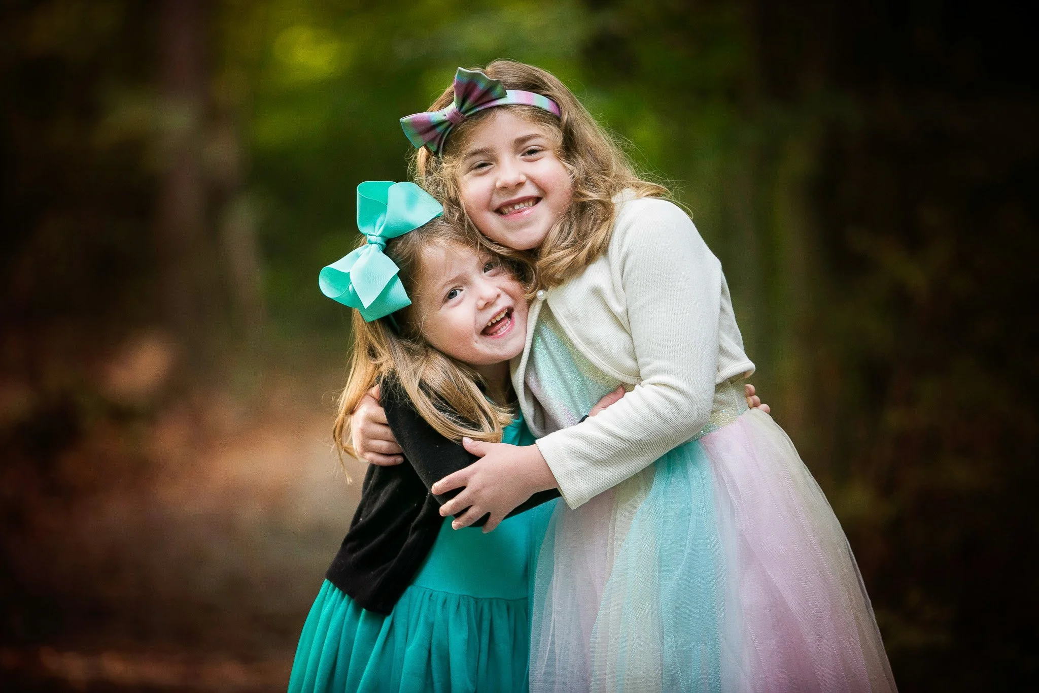 Two young girls hugging outdoors, smiling at the camera. One girl has long blonde hair with a large turquoise bow and wears a teal dress with a black cardigan. The other girl has curly blonde hair with a multicolored bow and wears a pastel-colored tu