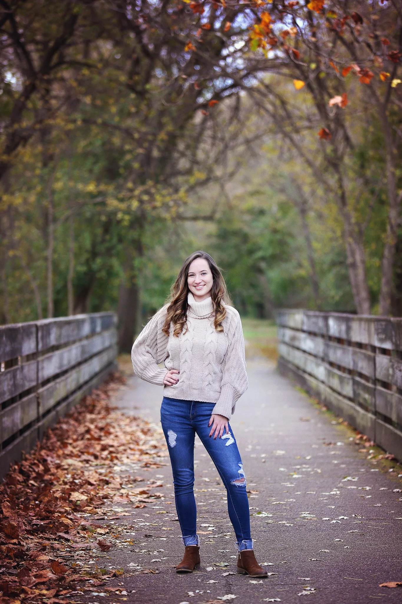 A young woman standing on a wooden bridge covered with fallen autumn leaves, surrounded by trees with changing leaves, smiling at the camera.