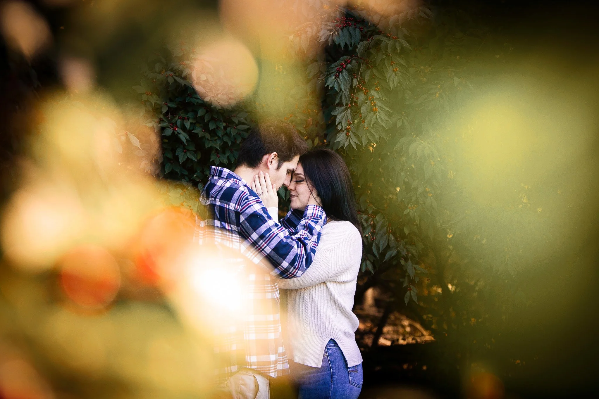 A young couple embracing outdoors surrounded by green foliage, with sunlight creating a soft glow.