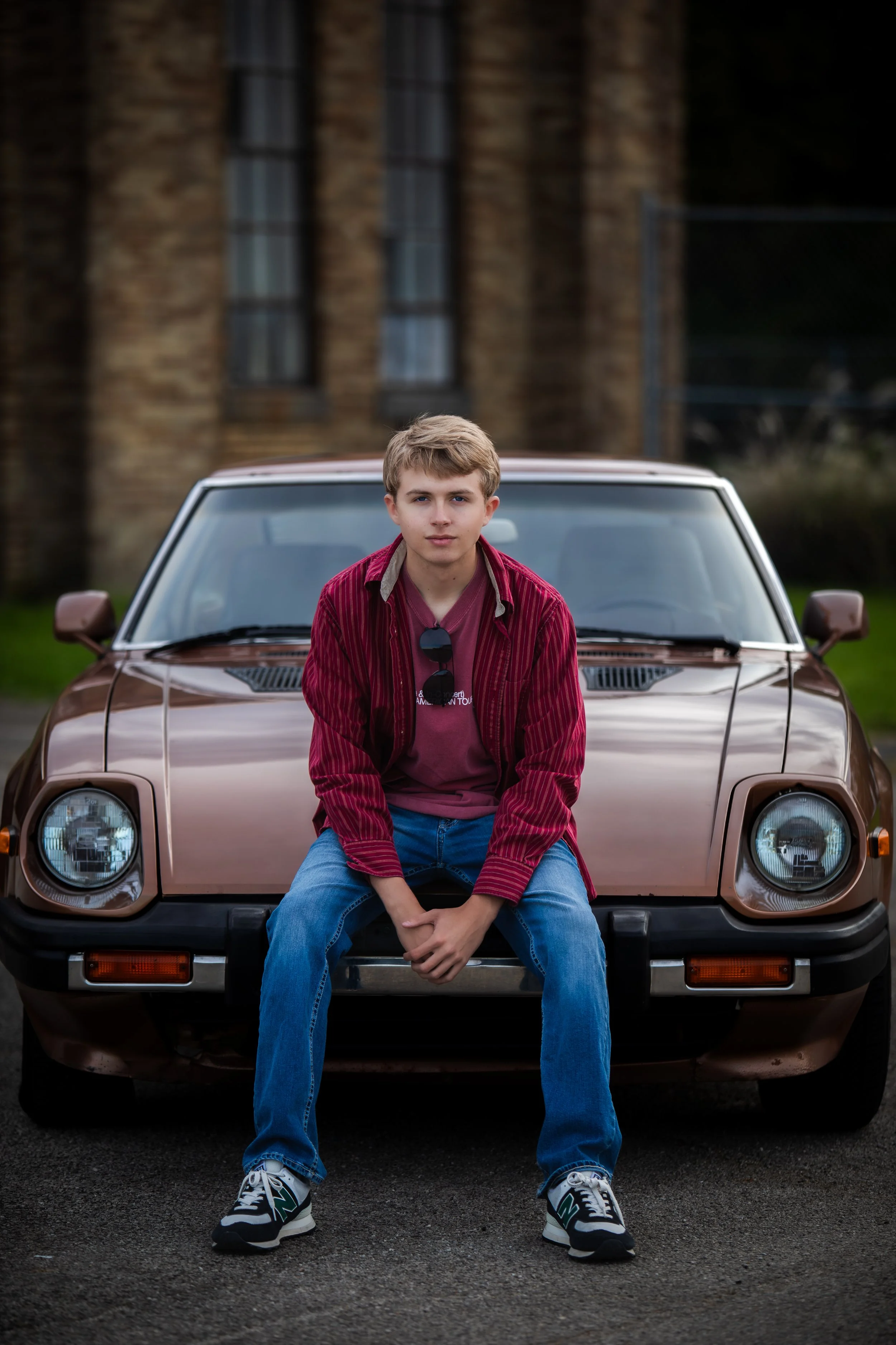 Young man sitting on the front bumper of a brown vintage car parked outdoors with a brick building in the background, wearing a red striped jacket, pink t-shirt, blue jeans, and sneakers.