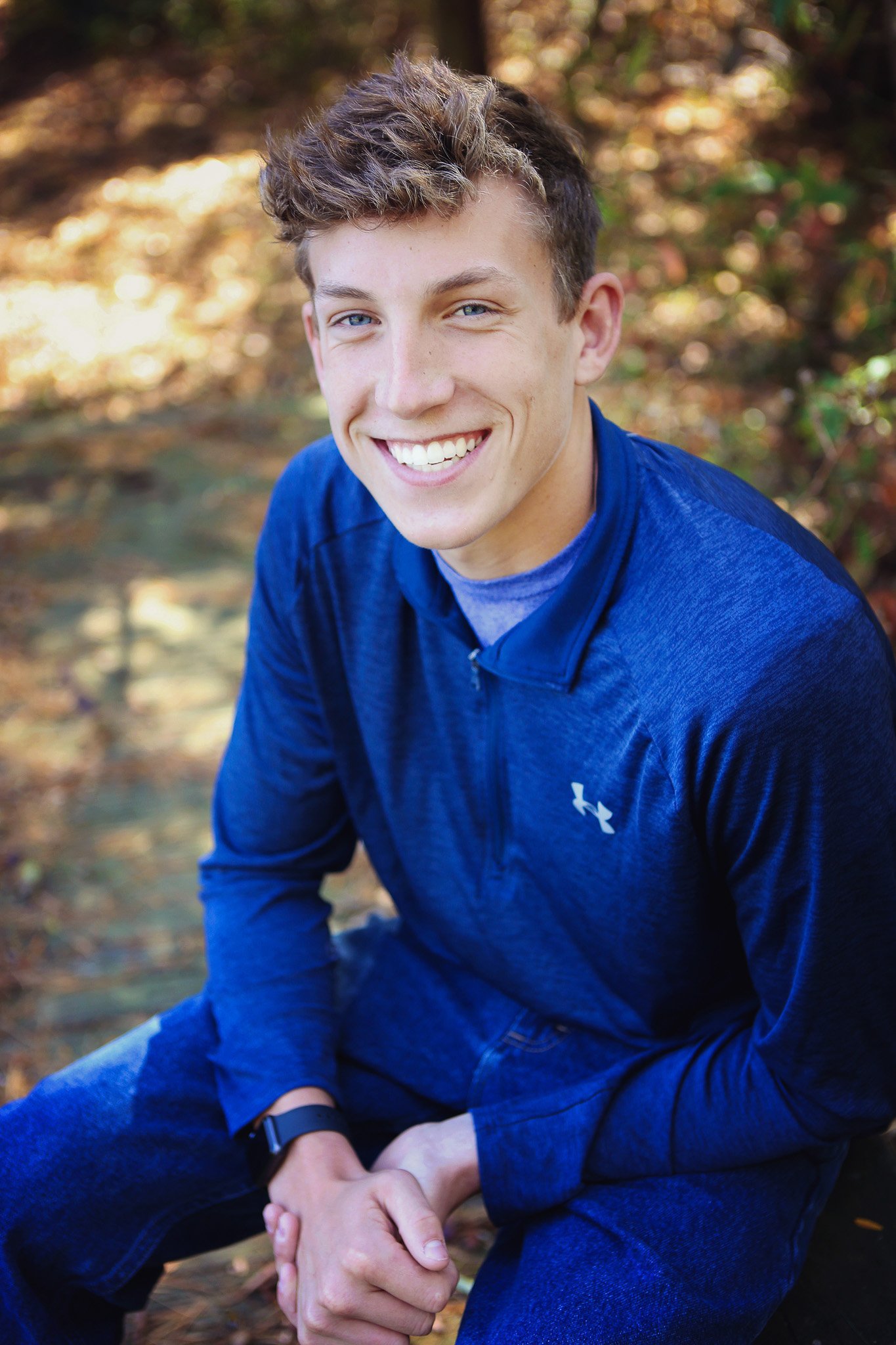 A young man with short, curly brown hair and blue eyes smiling at the camera. He is outdoors, sitting on a large rock in a natural setting with autumn foliage. He is wearing a blue Under Armour jacket, jeans, and a smartwatch on his left wrist.