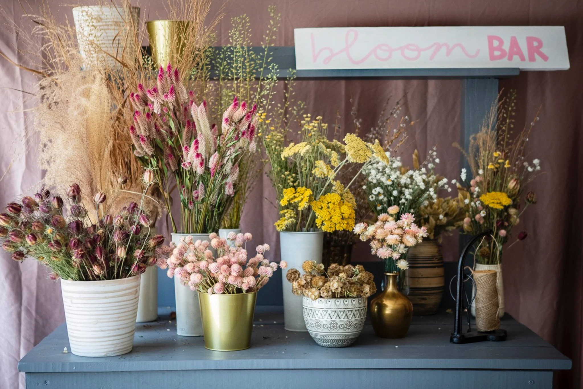A display of various colorful flowers in vases, with a handmade sign reading 'bloom BAR' above them, on a blue table against a purple backdrop.