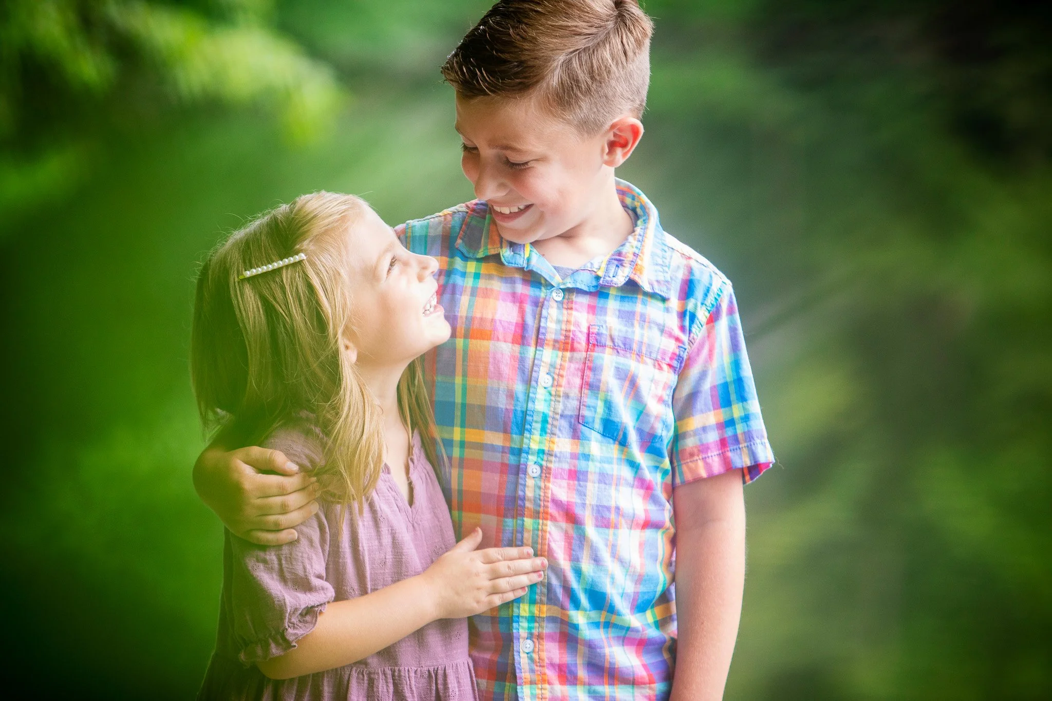 A young boy and girl smiling and looking at each other outdoors, with the boy's arm around the girl's shoulder.