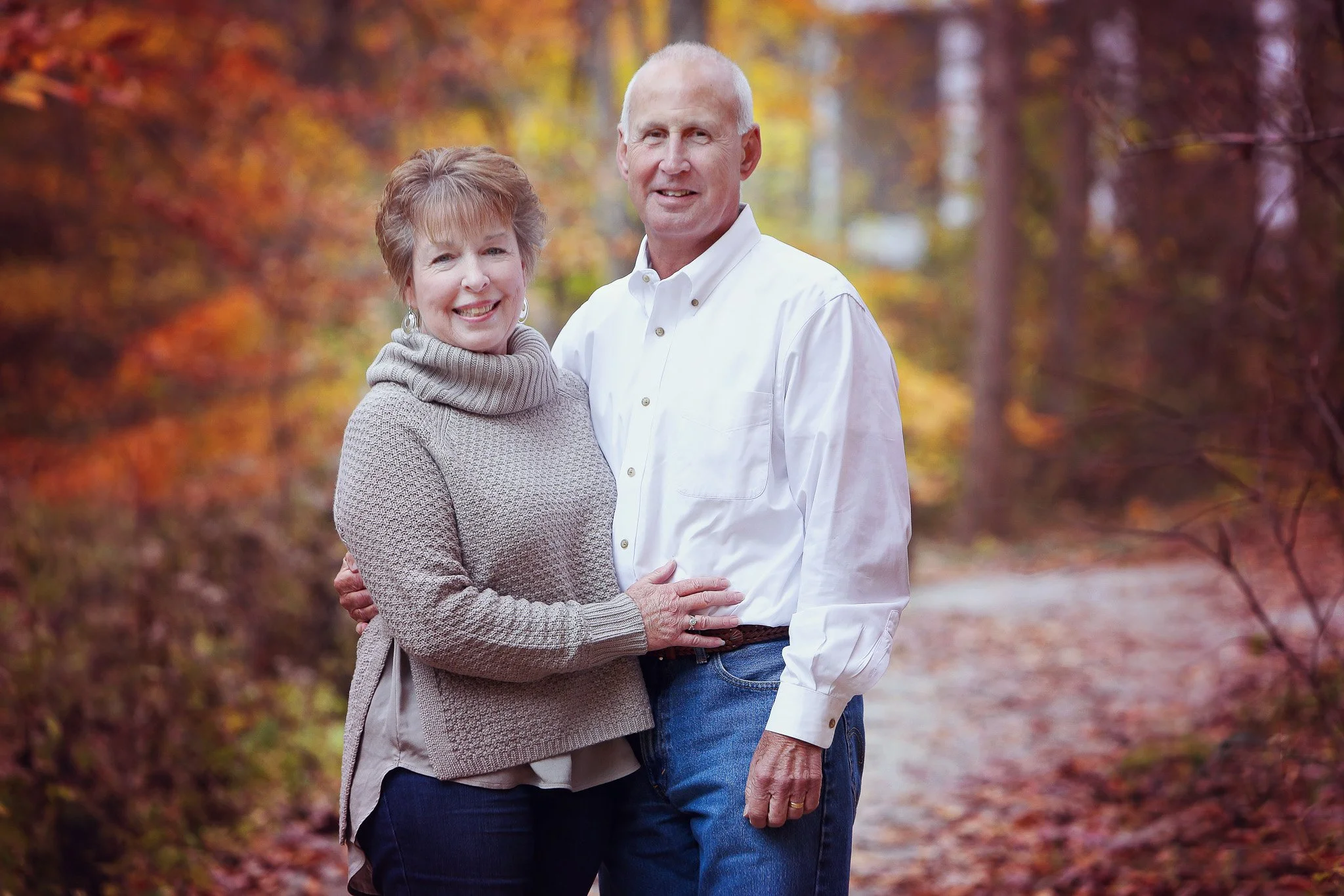 A happy older couple stands outdoors on a fall day, surrounded by colorful autumn trees. The woman has short, light brown hair and wears a beige turtleneck sweater and dark pants. The man is bald, wearing a white button-up shirt and jeans. They are s