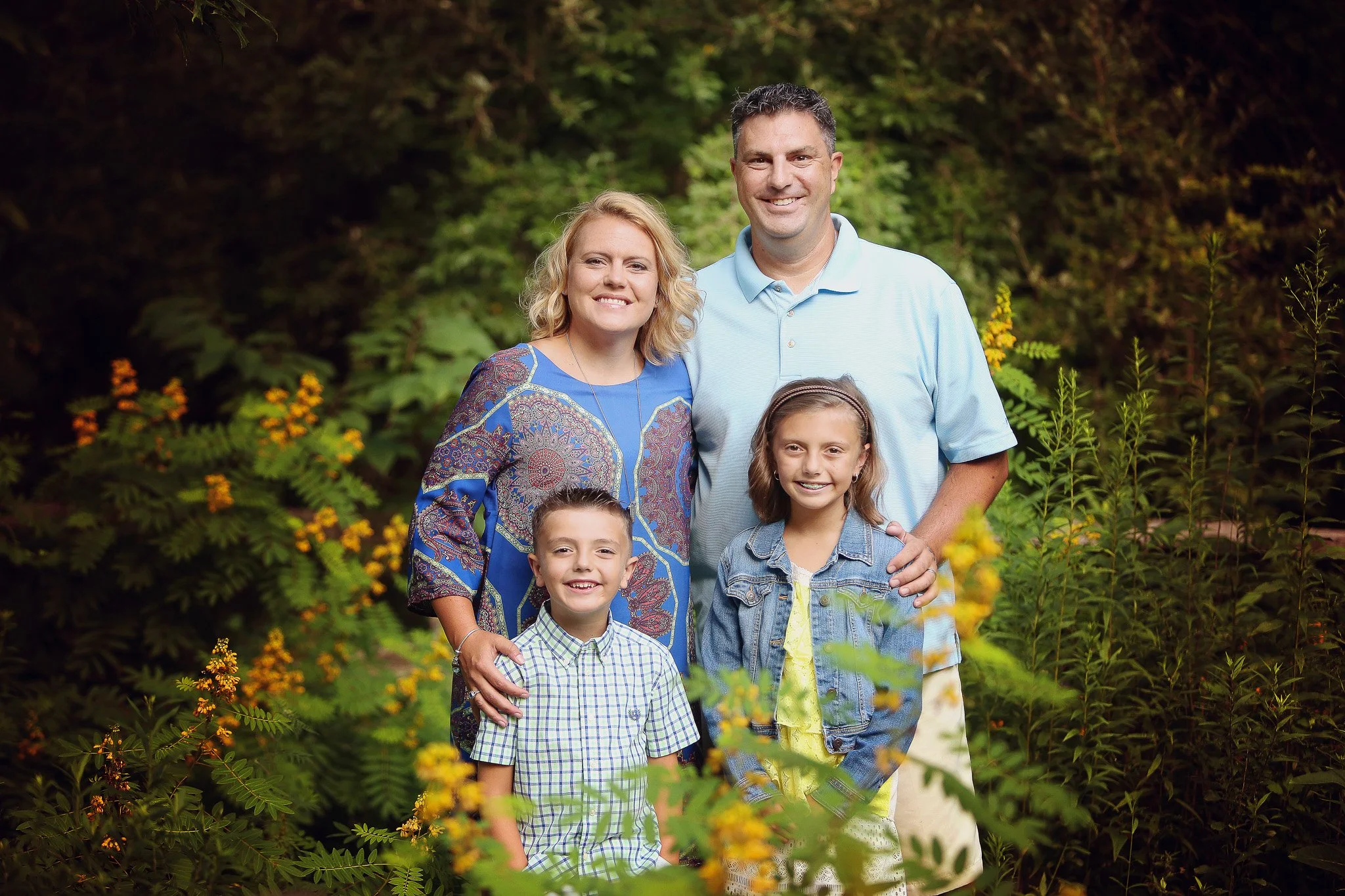 Family of four posing outdoors in a lush garden with green foliage and yellow flowers.