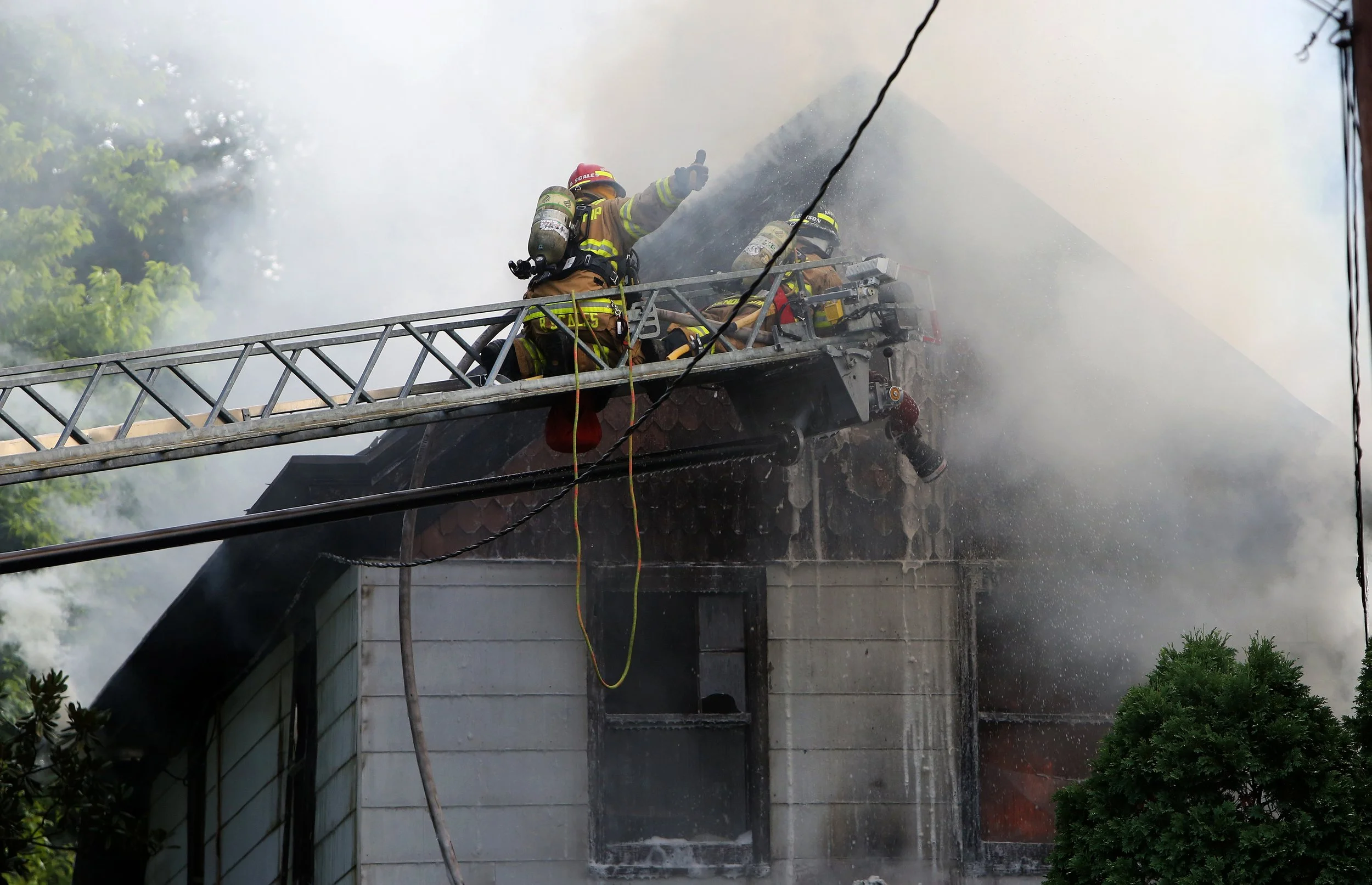 Firefighters on a ladder battling a house fire, with smoke billowing from the roof.