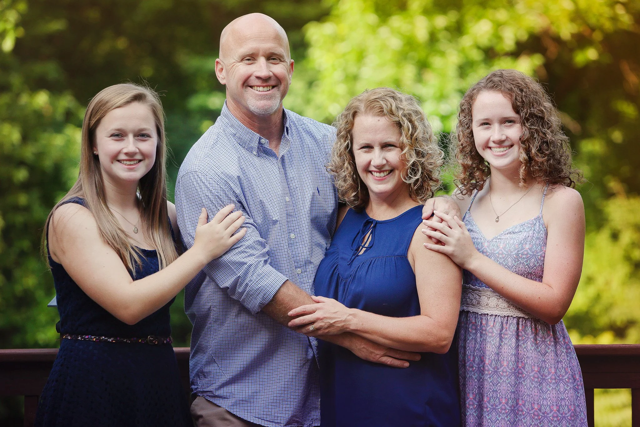 A family of two adults and two children smiling outdoors on a wooden deck with green trees in the background.