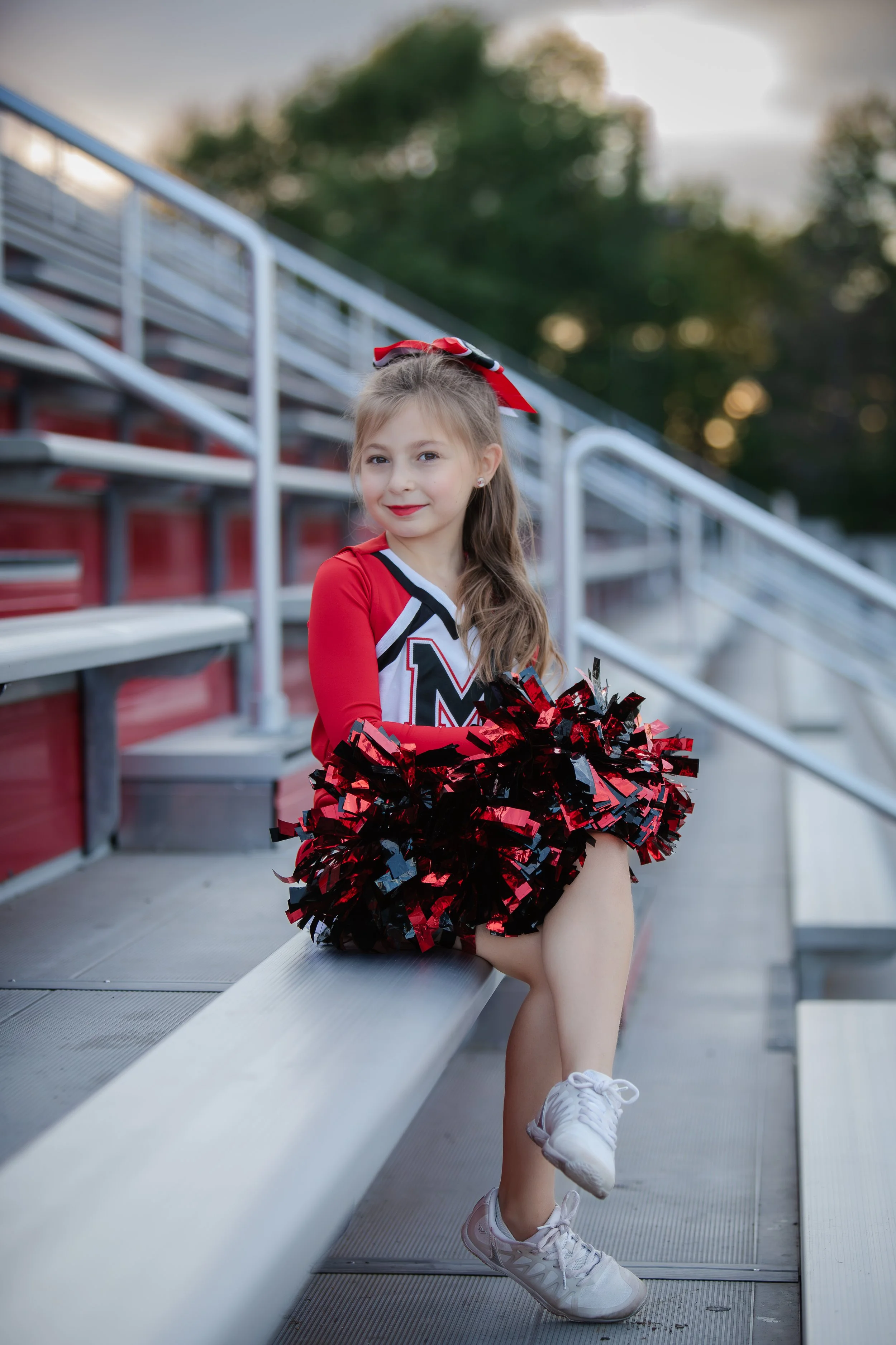 Young girl wearing a cheerleading uniform and pom-poms, sitting on stadium bleacher, outdoors during late afternoon or evening.