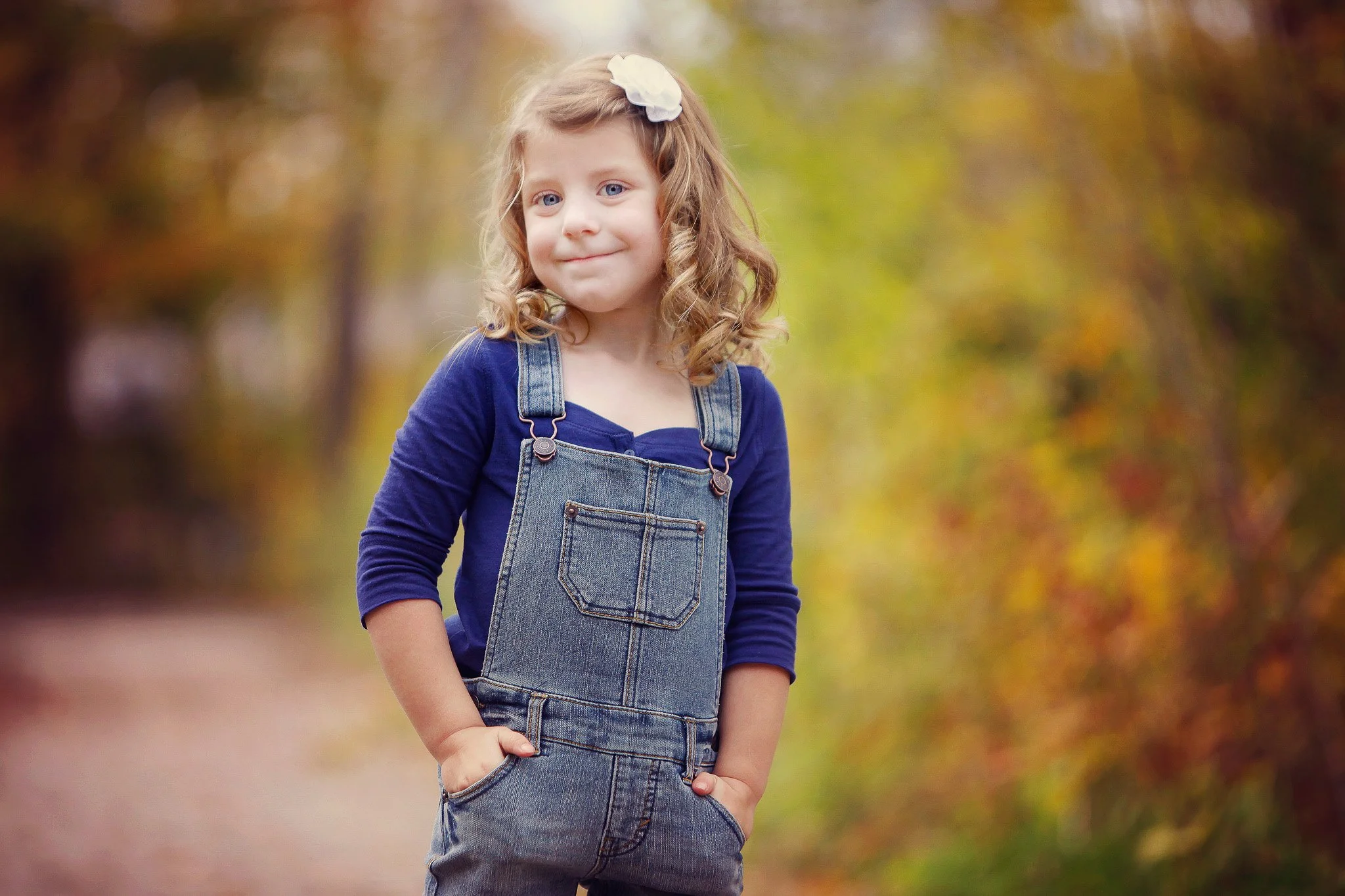 A young girl with curly blonde hair wearing a white hair bow, blue long sleeve shirt, and denim overalls standing outdoors on a path with trees showing fall foliage.