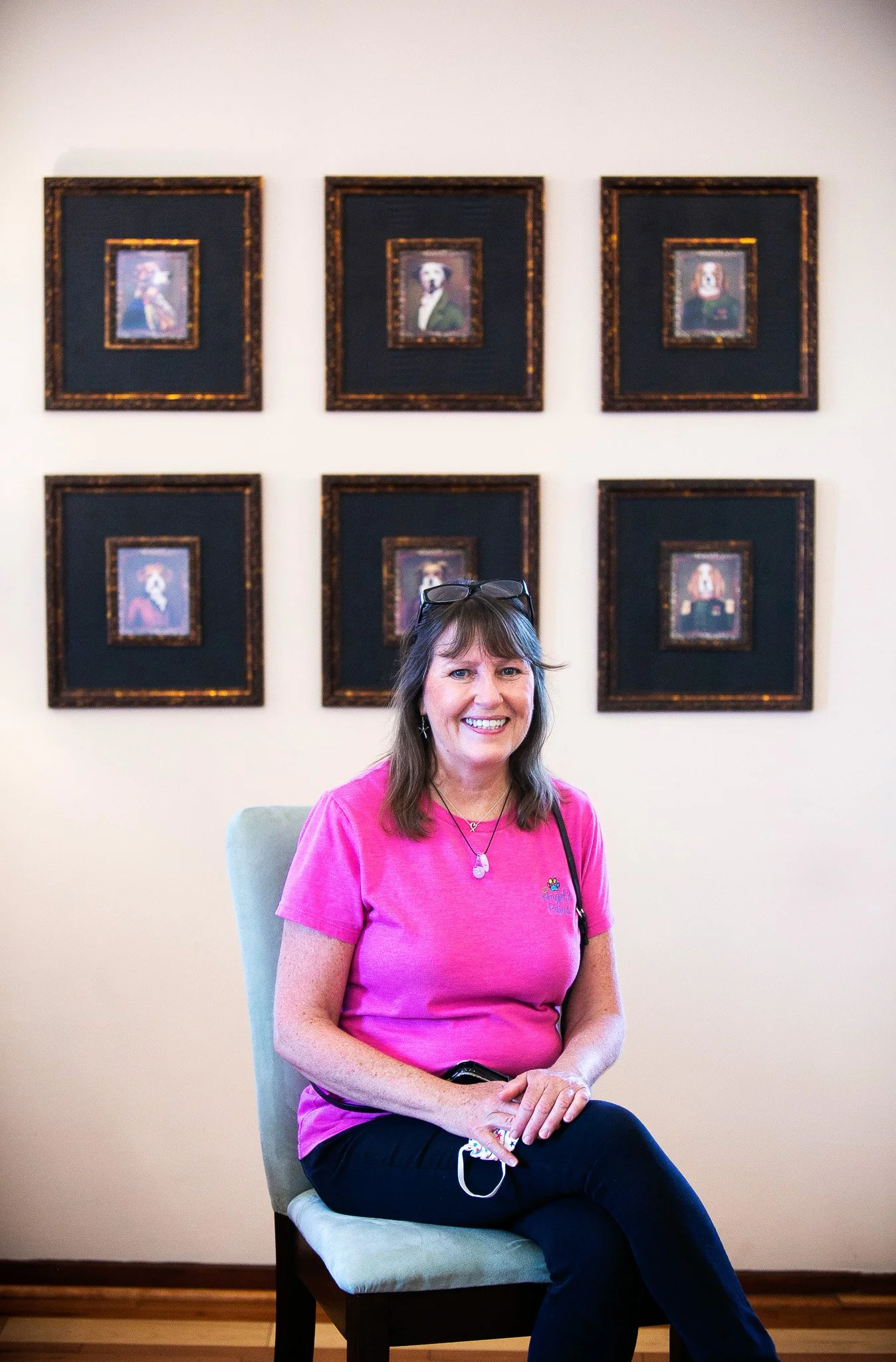 A woman with shoulder-length brown hair and glasses on her head, wearing a pink shirt and black pants, sitting on a light-colored chair in front of a wall with six framed pictures.
