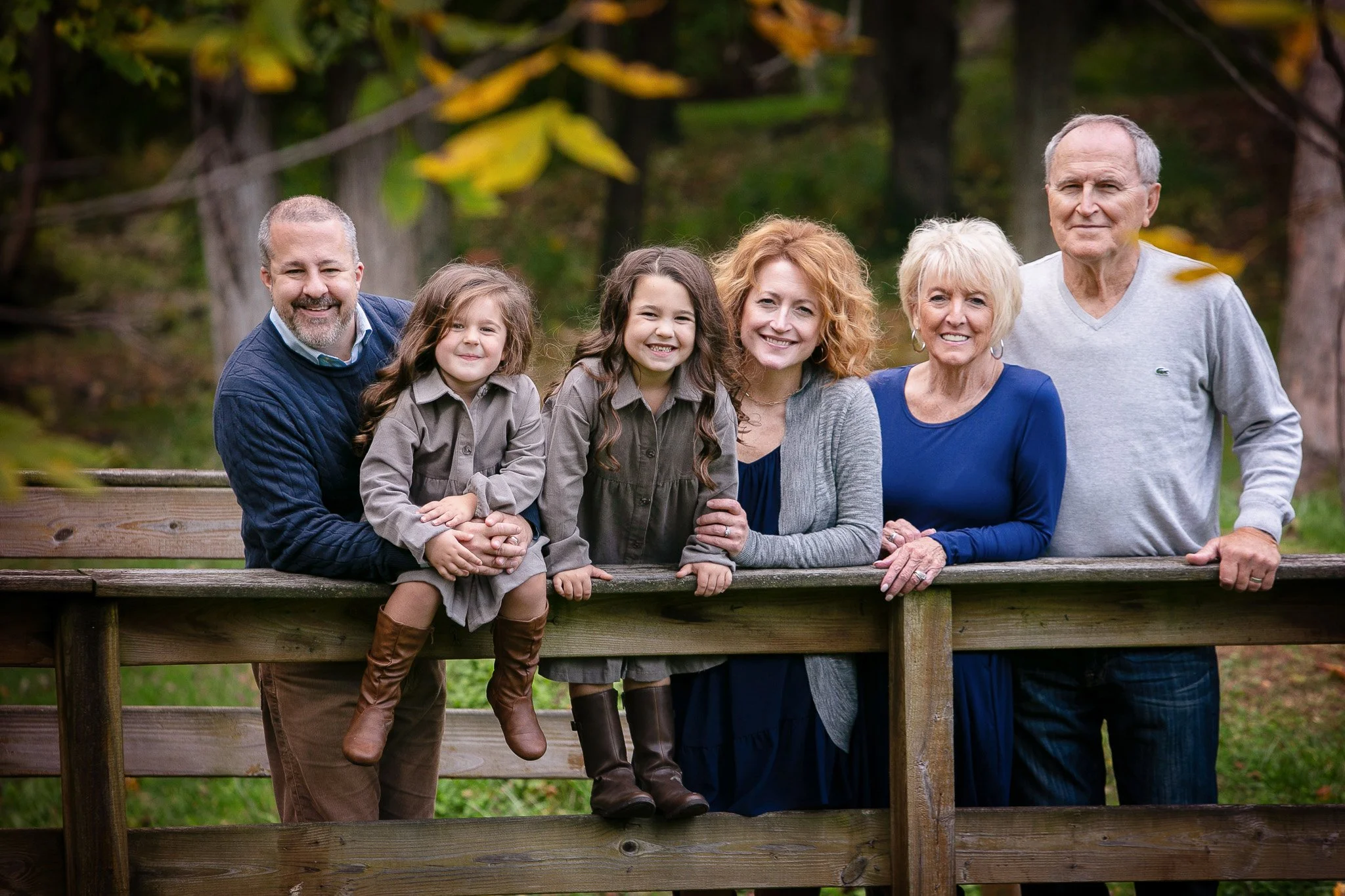 A multigenerational family of six standing outdoors behind a wooden fence, smiling. The group includes two elderly adults, two middle-aged adults, and two young girls. Trees with autumn leaves are in the background.