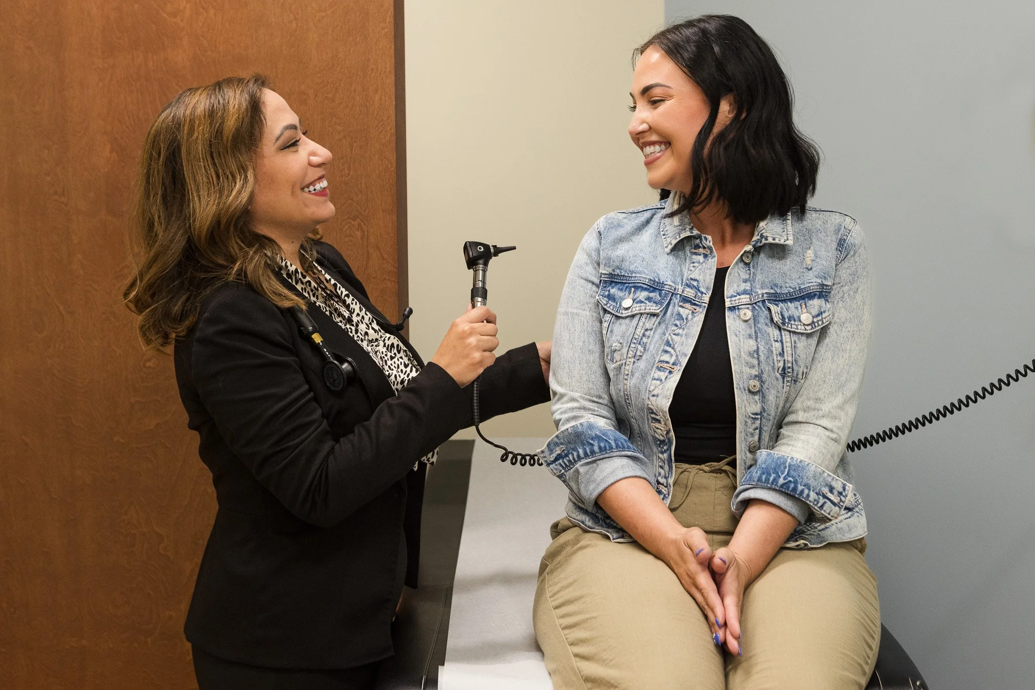 A healthcare professional holding a reflex hammer and smiling at a seated woman in a medical examination room.
