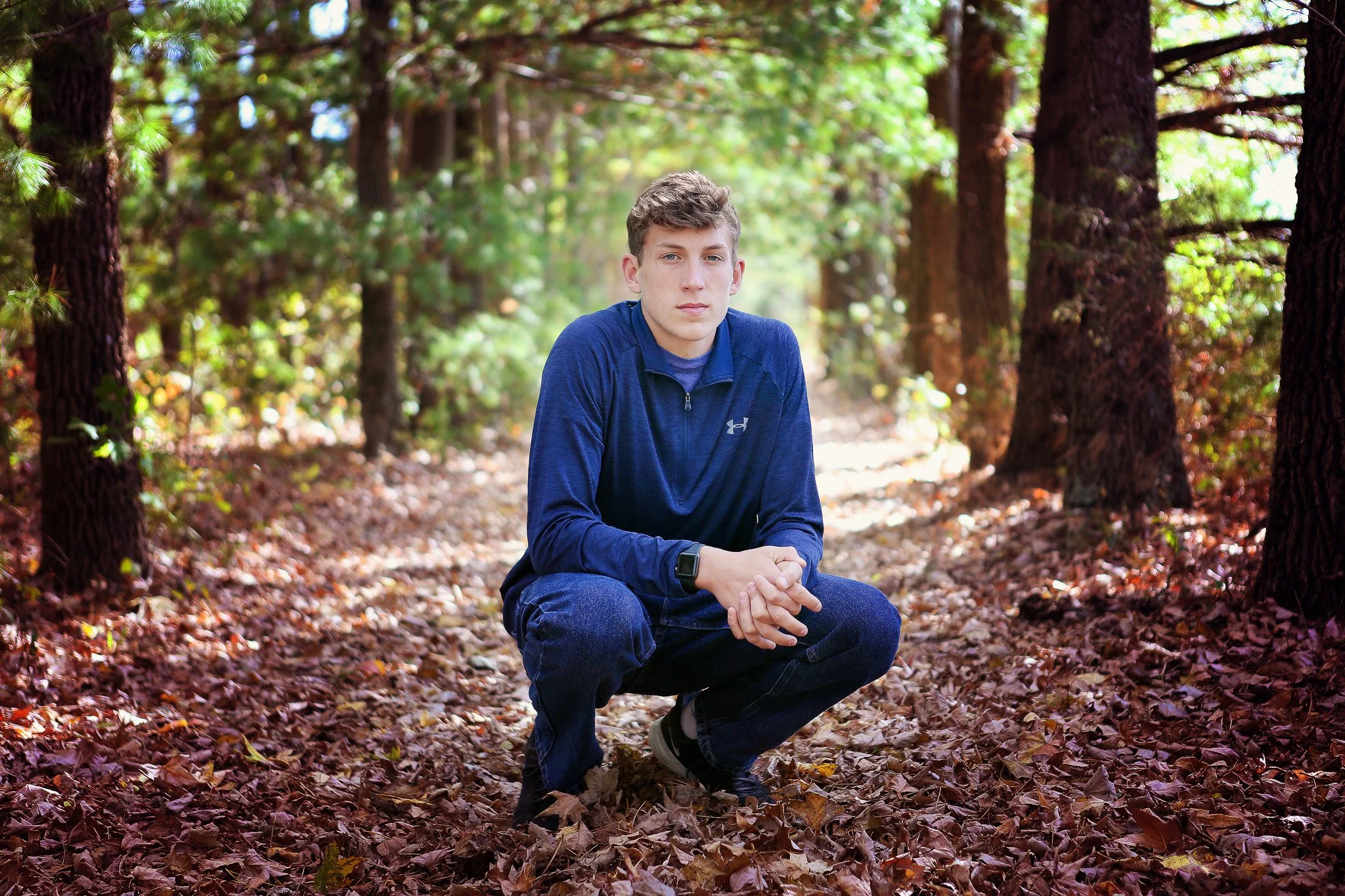 A young man crouching on a forest trail covered with fallen leaves.