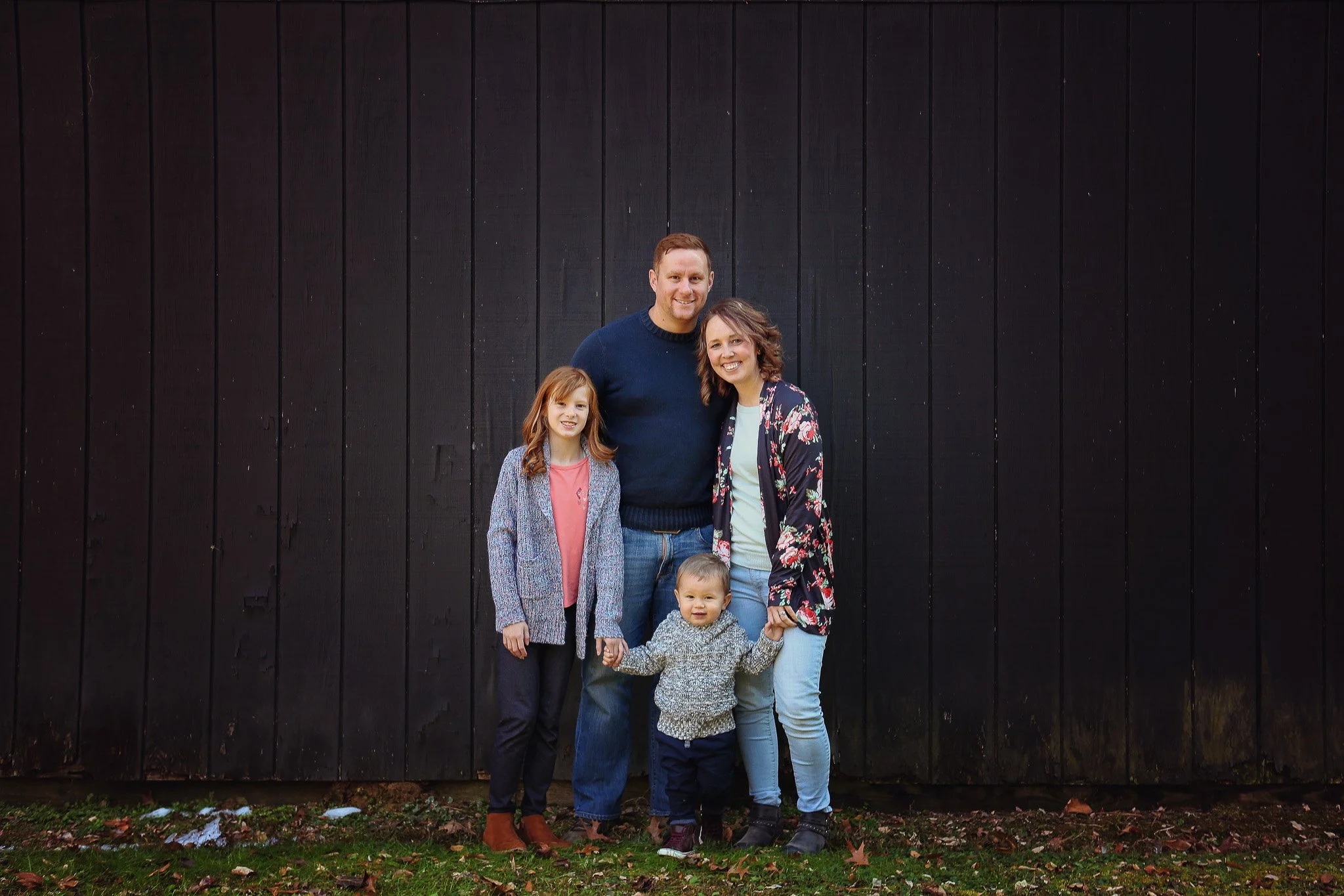 Family of five standing outdoors in front of a black wooden fence, holding hands, smiling.