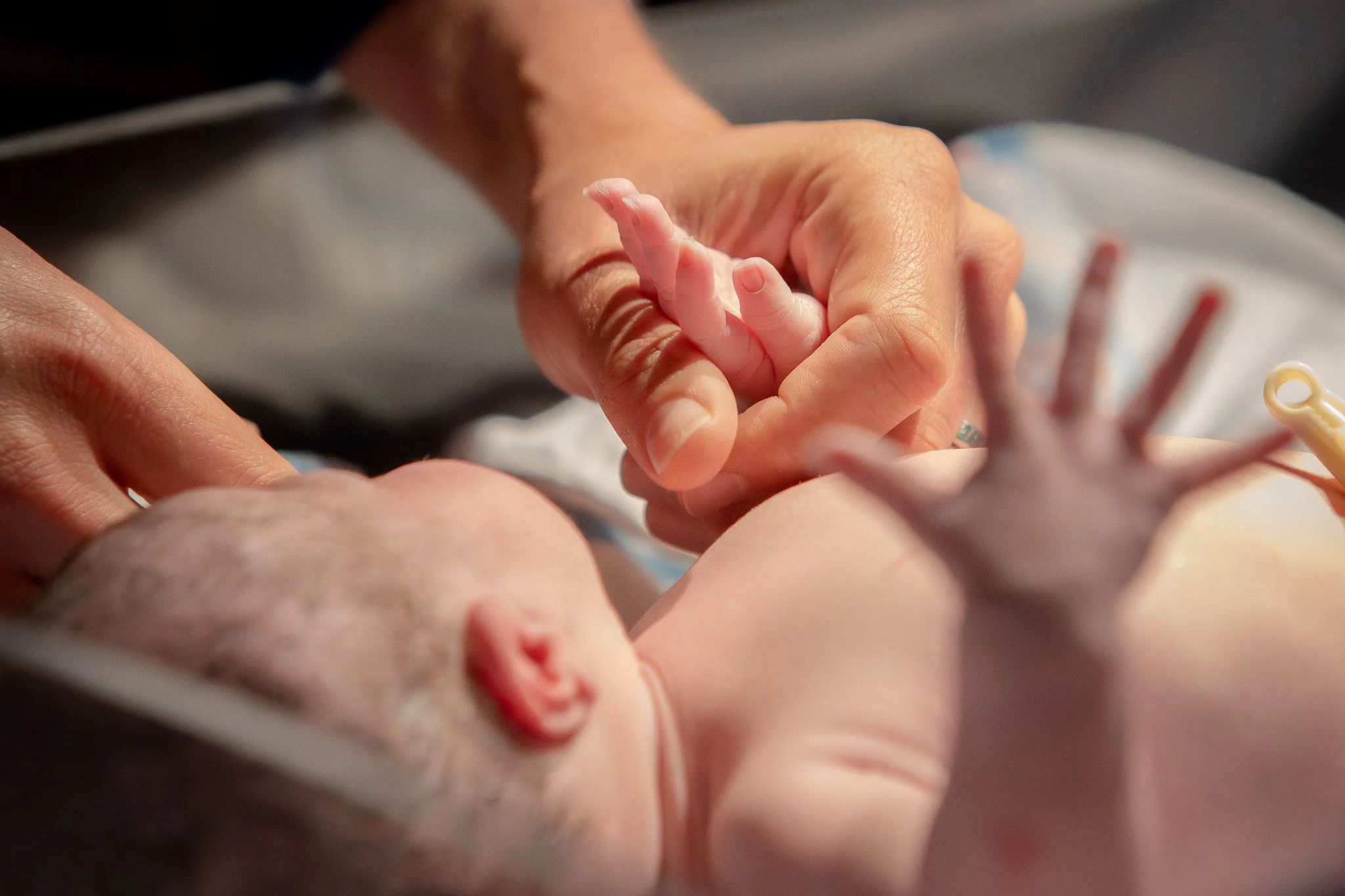 A newborn baby holding an adult's finger, showing close-up of hands and baby's head with a hospital setting background.