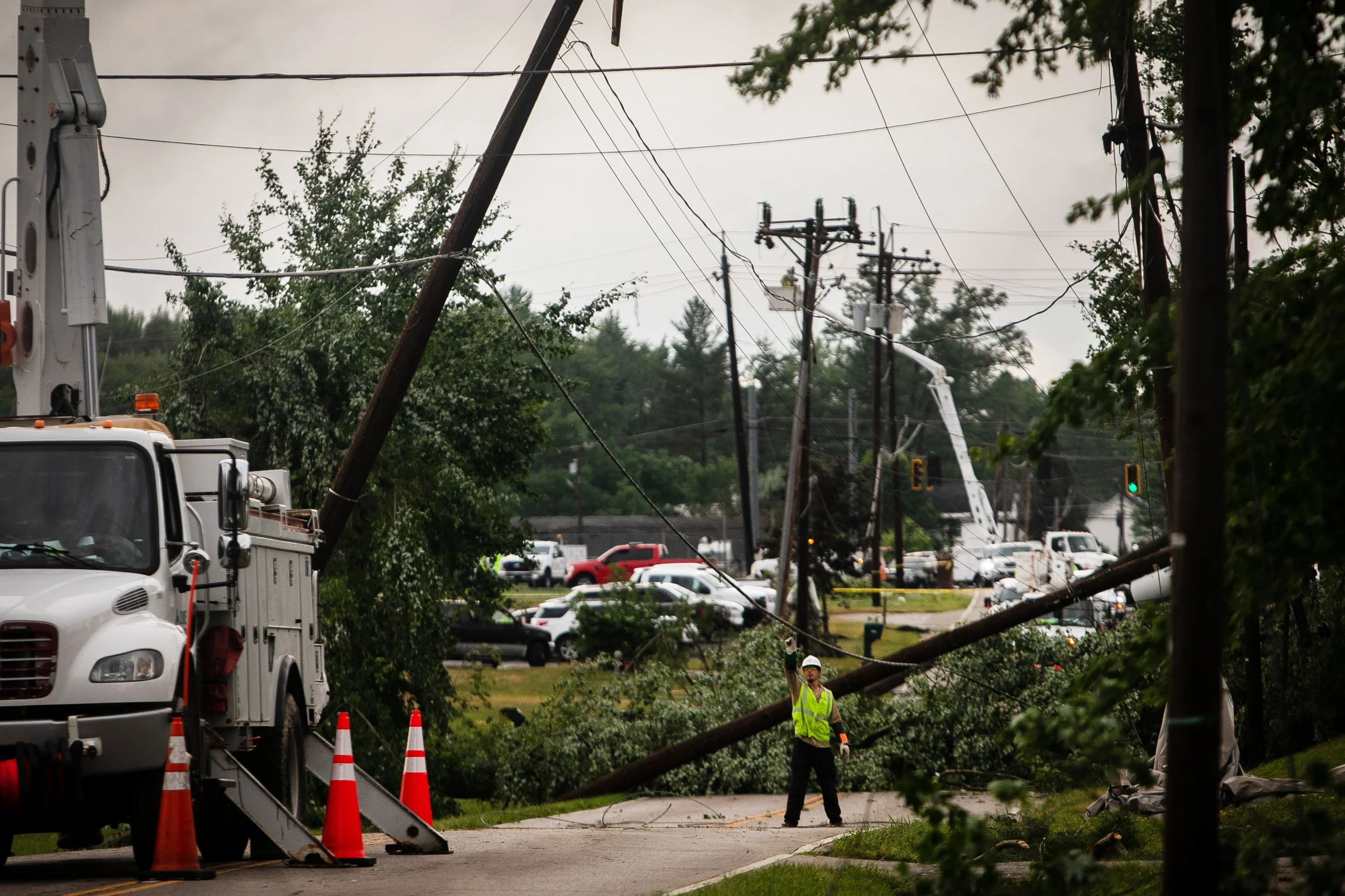 A utility worker wearing a yellow safety vest and helmet manages a fallen power line that is resting on the ground and leaning on a tree, caused by a storm or accident, with traffic and utility poles in the background.