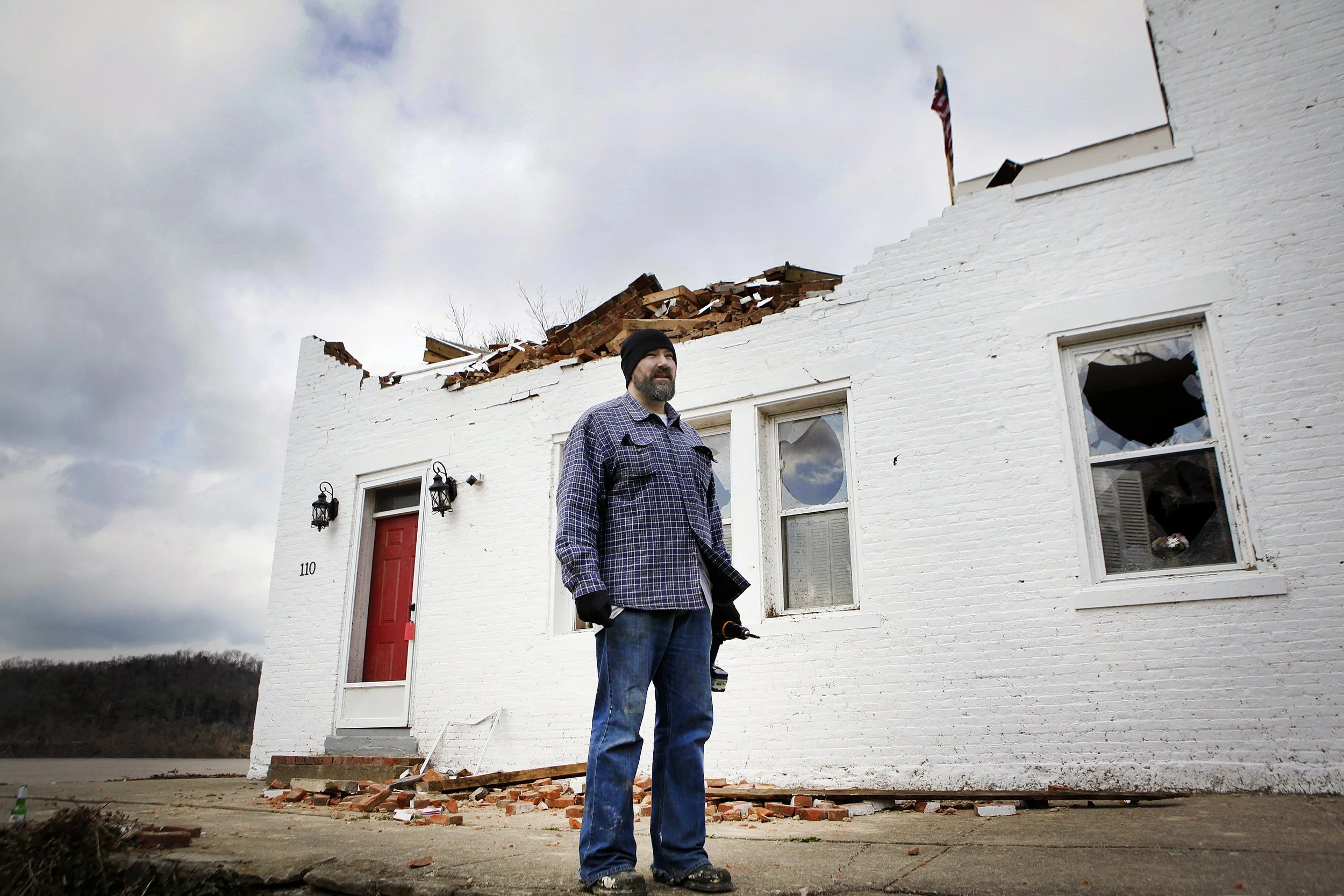 A man in a checkered shirt and beanie standing outside a partially demolished white brick building with a damaged roof and broken windows.
