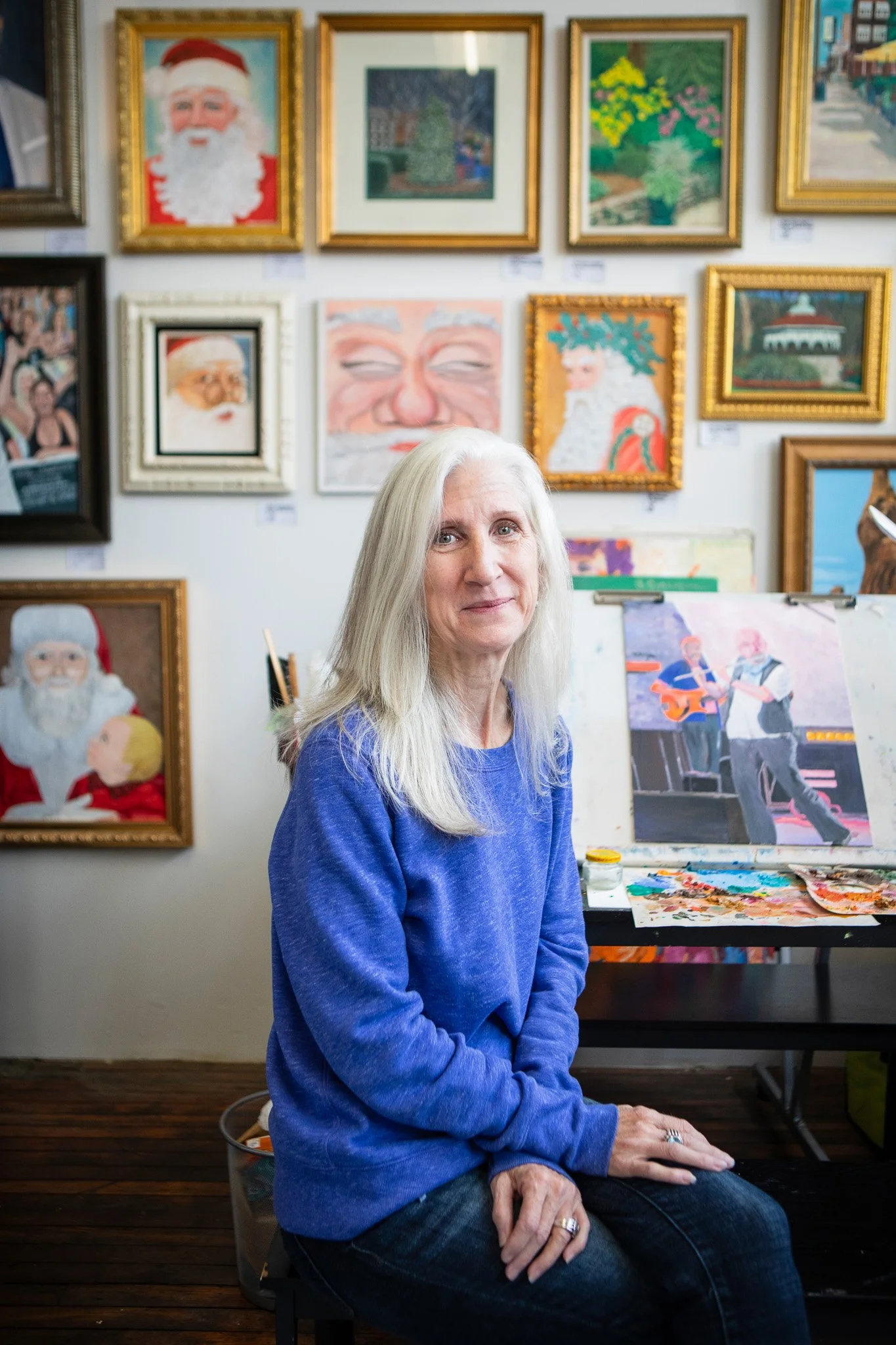 An elderly woman with long white hair wearing a blue sweatshirt sitting in an art studio. Behind her, framed paintings include a Santa Claus theme, faces, flowers, and landscapes.