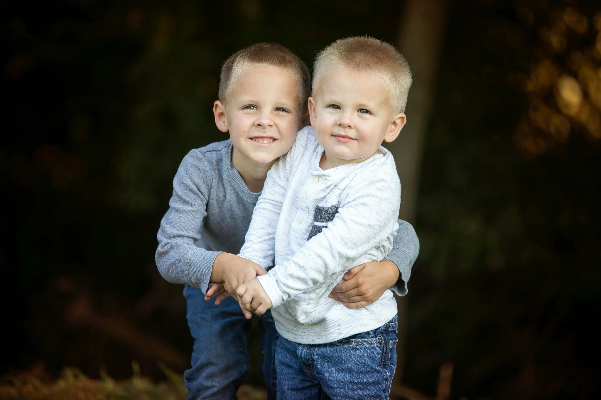 Two young boys with blonde hair, wearing long-sleeve shirts and jeans, are hugging each other outdoors with trees in the background.